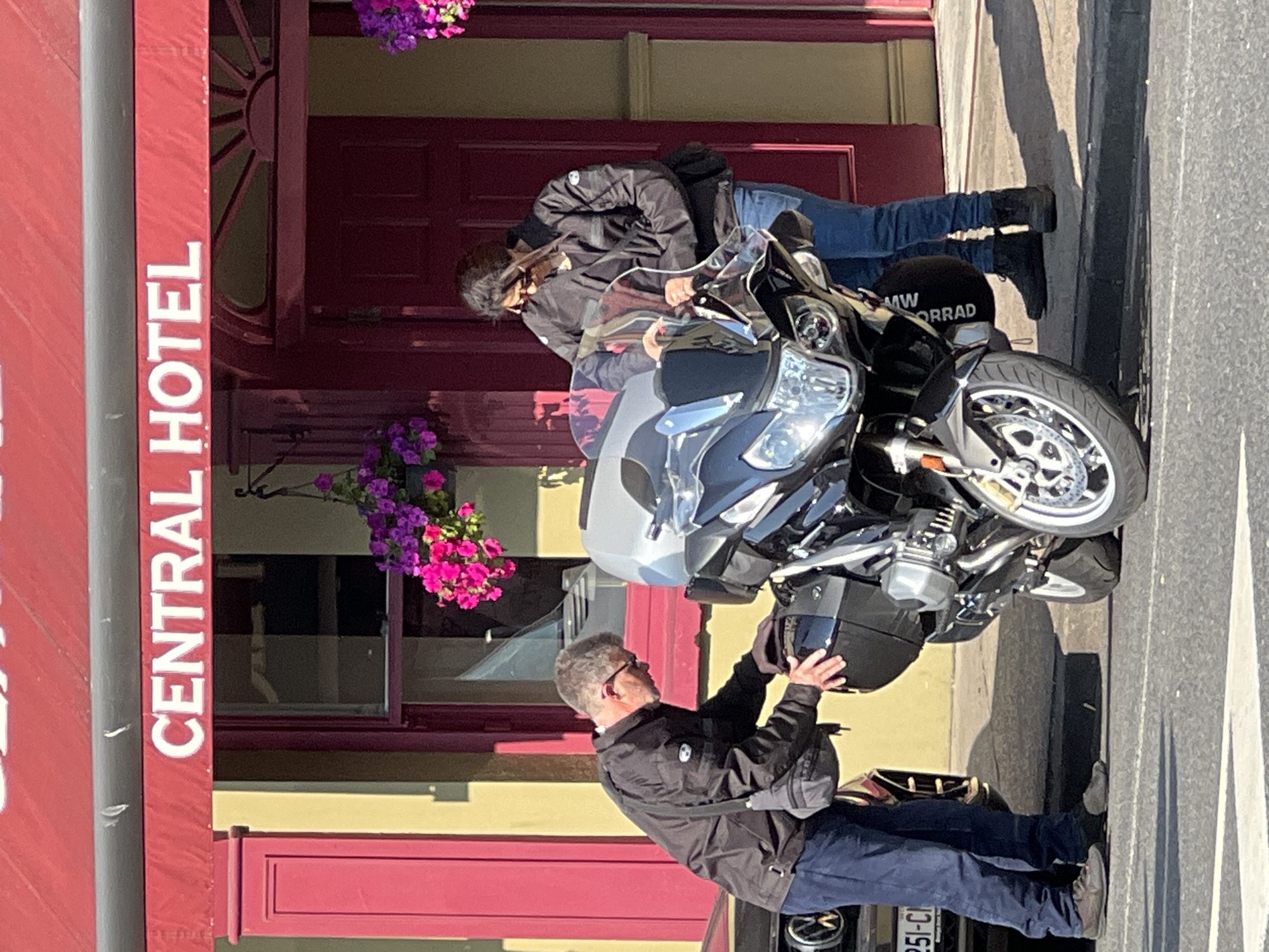 Two individuals, a man and a woman, are standing beside a black motorcycle in front of a building with a red awning displaying the words "CENTRAL HOTEL" in white lettering. The man, wearing a black jacket and blue trousers, is bent over, attending to the motorcycle's storage compartment. The woman, also in a black jacket and blue jeans, is looking at the motorcycle, her hands near its windshield. The building appears to be made of yellow brick and has red doors and window frames. Potted plants with pink and purple flowers hang near the entrance. The scene takes place on a paved street in what appears to be the city of Donegal, Ireland. A portion of a car is visible to the left of the motorcycle.