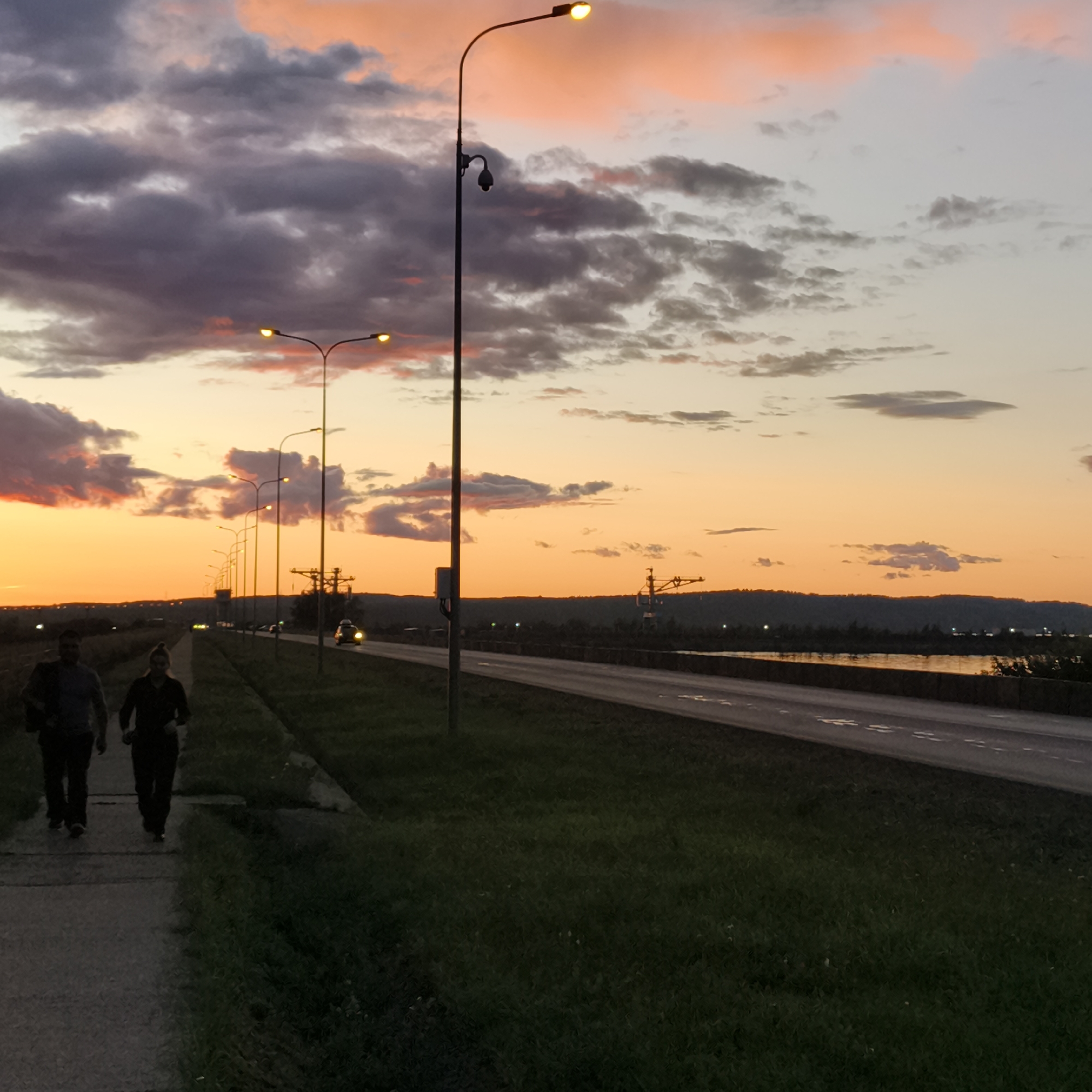 Two people, a man and a woman, are walking on a paved path alongside a grassy area. The man is wearing a dark jacket and pants, and the woman is wearing dark clothing. They are walking away from the camera. A road is to their right, with several cars visible, some with their headlights on. A line of streetlights runs along the road. In the background, there is a body of water reflecting the orange and pink hues of the sunset. Hills are visible on the horizon. The sky is filled with dramatic clouds, with streaks of orange, pink, and dark gray. The overall impression is of a twilight scene in an urban or suburban environment. The image appears to be taken in Chaikovsky, Russia, given the context provided.