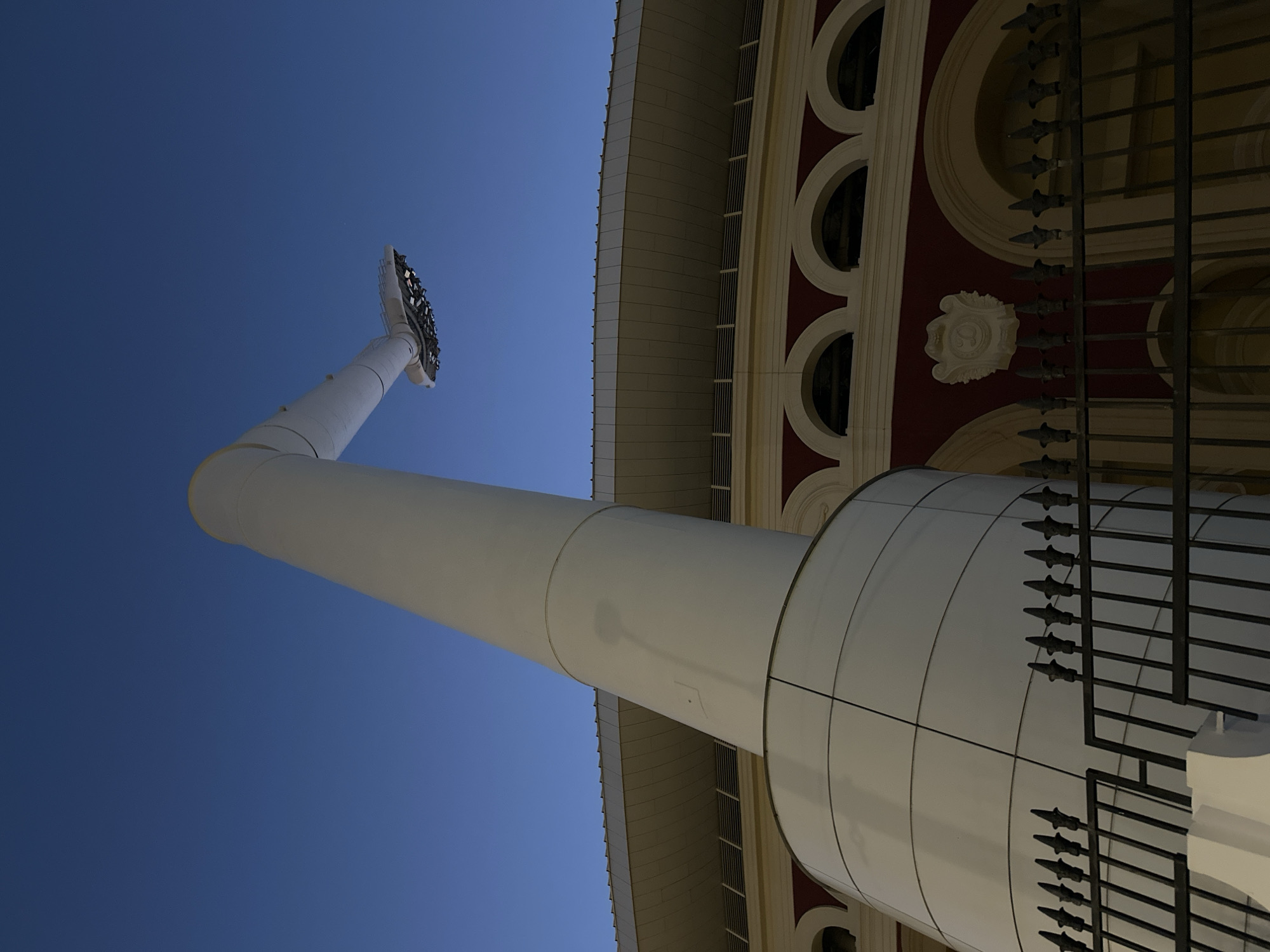 A low-angle, wide shot captures a portion of a large, modern stadium structure under a clear, deep blue twilight sky. Dominating the left side of the frame is a massive, white, cylindrical pillar that tapers as it extends upwards. This pillar bends at its top, extending horizontally to support a large, circular lighting fixture with visible bulbs. The stadium's upper structure is visible behind the pillar, featuring a curved, beige roof with a ribbed underside and a darker band below it. Beneath this, a section of the stadium's facade is visible, painted a deep red and adorned with a row of arched openings with pale yellow infills. Ornate decorative elements are present on the red facade. In the foreground, a black, ornate metal fence runs horizontally across the lower portion of the image, partially obscuring the lower sections of the stadium. The overall lighting suggests it is either dusk or dawn, with artificial lights on the stadium and the floodlight beginning to illuminate. There are no people or animals present in the image.