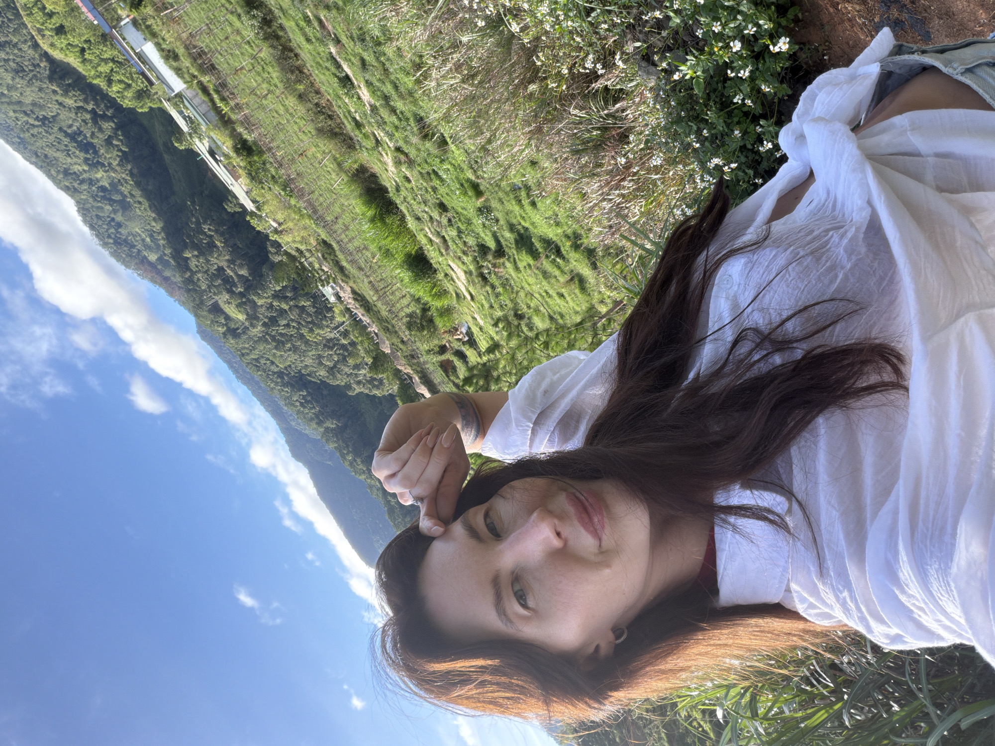 A woman with long, dark brown hair poses for a selfie in a hilly, green landscape under a bright blue sky with scattered clouds. She is wearing a loose-fitting white shirt and denim shorts. Her right hand is raised to her forehead, and she is looking at the camera with a slight smile. The background features lush, terraced hillsides with scattered buildings and vegetation. The exact location in Klong Klanh, Vietnam, is not discernible from the image itself, but the environment suggests a rural, mountainous area.