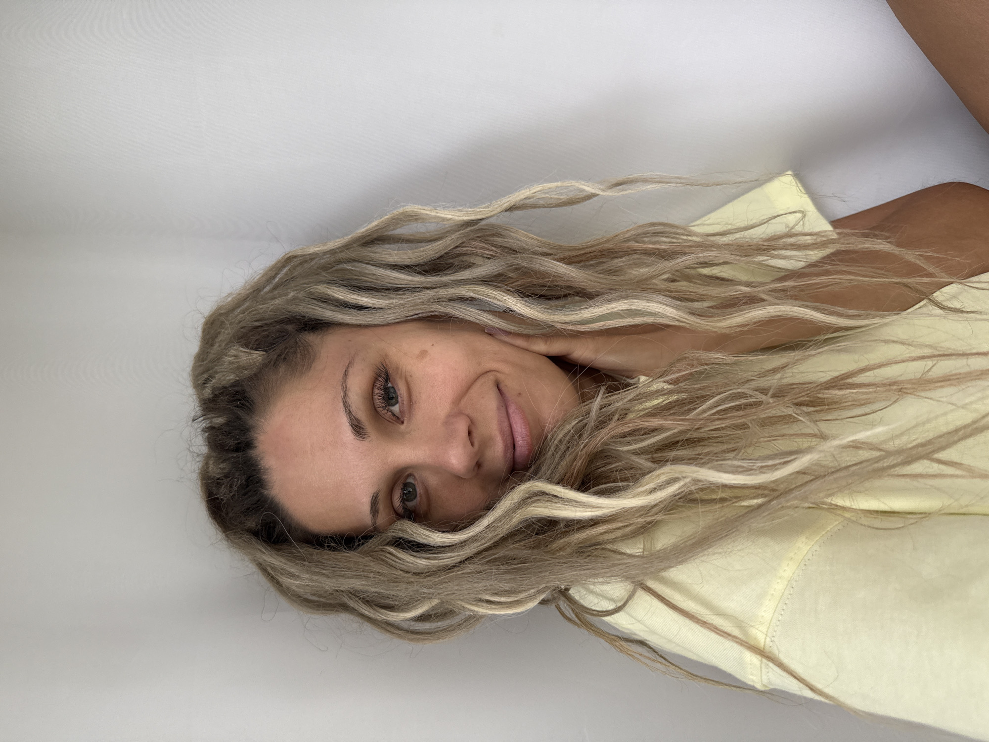 A medium shot captures a woman with blonde, wavy hair and light skin, wearing a yellow t-shirt. She is positioned against a white wall, with her right hand resting on her cheek. Her eyes are open, and she is smiling slightly. The lighting is natural, creating soft shadows. The scene is likely indoors, given the plain wall background. There is no indication of the city of Rusanovo, Russia in this image.