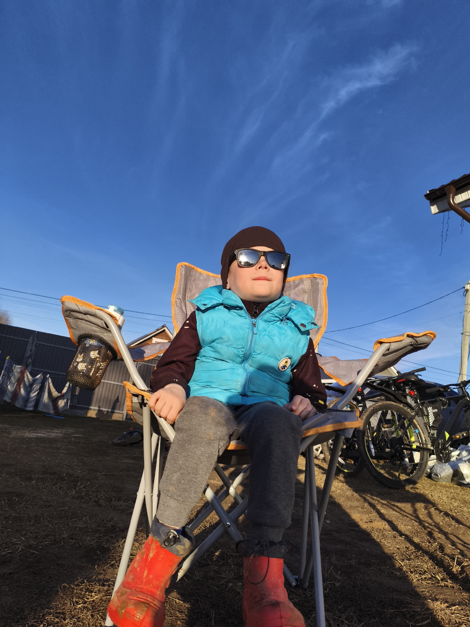 A young child, approximately 5-10 years old, is seated in a grey and orange folding camp chair outdoors. The child wears a dark beanie, black sunglasses, a bright blue puffer vest over a dark long-sleeved shirt, grey sweatpants, and distinctive red, mud-splattered boots. The child looks slightly upwards and to the right, appearing relaxed.
The setting is an outdoor area, likely a backyard or rural space in Русский Вожой, Russia. The ground is covered with dry grass and dirt, showing long shadows cast by the golden hour sunlight, indicating late afternoon or early evening. The sky is a clear, vibrant blue with a few wispy clouds. In the background, a fence, parts of a building's roofline with a gutter, utility poles, and at least two bicycles are visible. The folding chair has a mesh cup holder attached to its left armrest. No legible text is visible in the image.