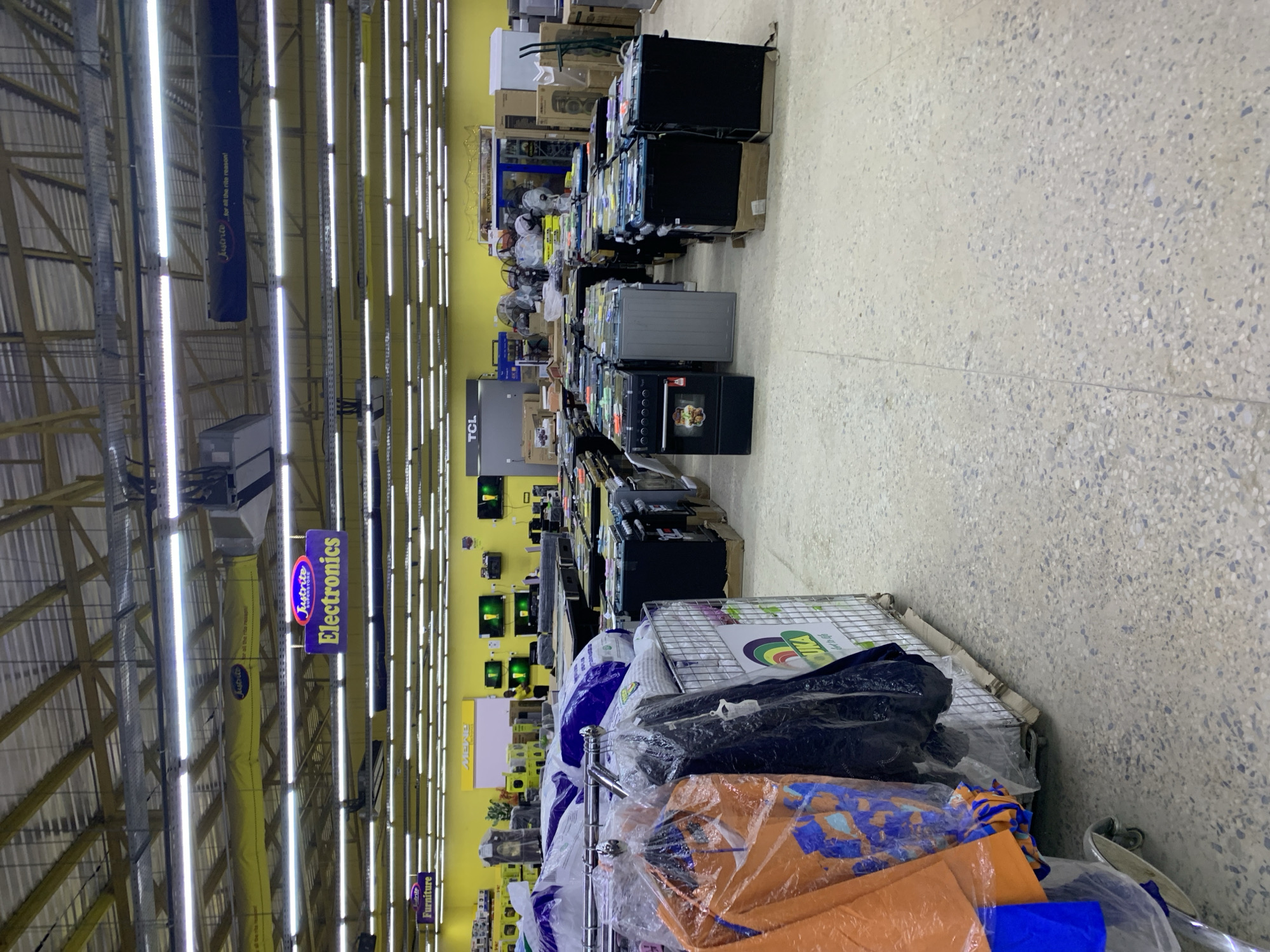 The image shows a retail store, likely an electronics store, with aisles of merchandise. The perspective is from a low angle, looking down an aisle filled with appliances such as ovens, televisions, and other electronic devices. Many of the items are in their original packaging, stacked on shelves or displayed on the floor.
In the foreground, there are several large items wrapped in plastic, possibly bedding or textiles, on a display rack. The store is brightly lit by rows of fluorescent lights on the ceiling. The walls are painted yellow, and there are signs hanging from the ceiling indicating different sections of the store. One sign clearly reads "Electronics" with the "Jutrite Superstore" logo. Another sign in the distance appears to say "Furniture."
The floor is a speckled terrazzo pattern. The overall impression is that of a busy retail environment, though no people are visible in the frame. The lighting suggests it is daytime, and the indoor setting makes weather conditions irrelevant. The arrangement of products suggests a sale or a restocking process, with items ready for display or purchase. The abundance of electronics and the prominent "Electronics" sign strongly indicate the nature of the store.