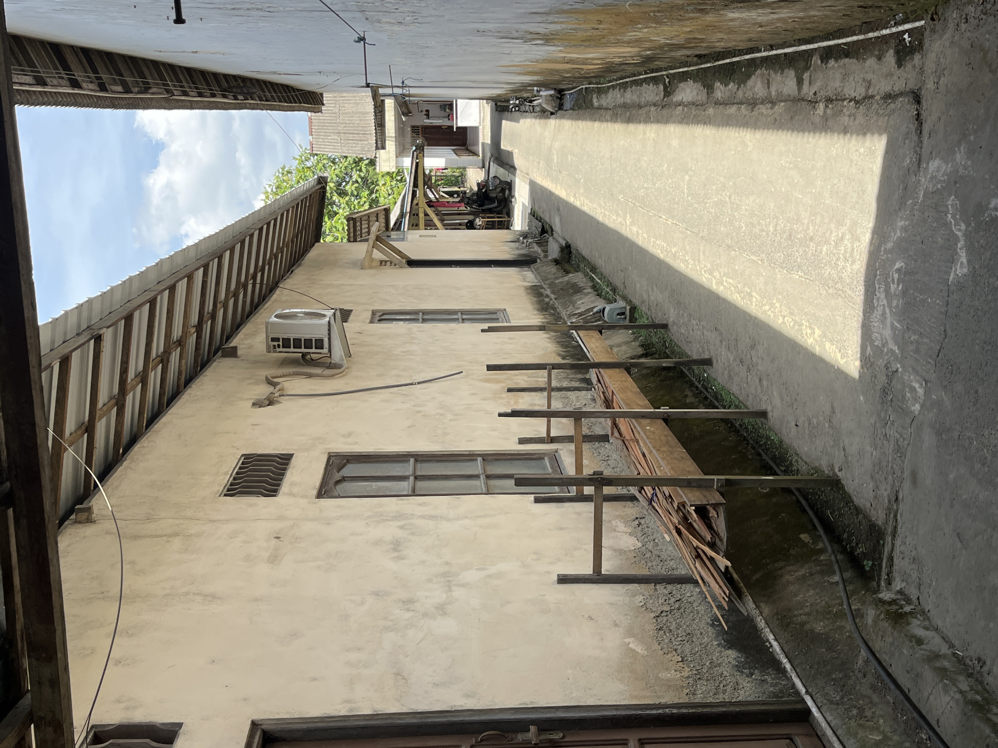 The image depicts a narrow alleyway in the city of Sejahtera, Indonesia, flanked by two buildings. The building on the left is made of textured, light-colored plaster and features a wooden door, a barred window, and a rectangular window with multiple panes. An air conditioning unit is mounted on the exterior wall. A metal railing system with wooden planks is positioned along the base of this building, with a pile of lumber resting on it. The alley floor is paved with concrete, and a drainage channel runs alongside the building on the left. The building on the right is also plastered, with visible discoloration and some debris along its base. The alley extends into the distance, where other buildings and greenery are visible. The sky above is partly cloudy. There are no people clearly visible in the scene.