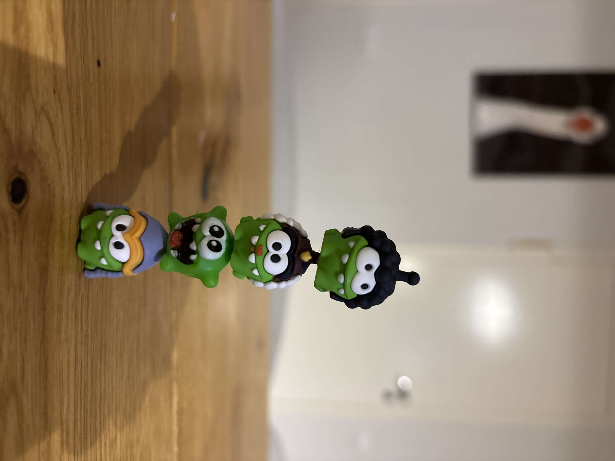 A straight, eye-level shot shows four green, cartoon monster figurines lined up on a wooden surface. The first figurine is a light green with a blue and yellow hat. The second is a darker green with an open mouth. The third is a lighter green with a white and brown hairstyle. The fourth is a darker green with black, curly hair. The background is a white wall with a blurry black and white picture and a doorknob. The setting appears to be indoors. There is no verifiable evidence of the city of Chaikovsky, Russia.