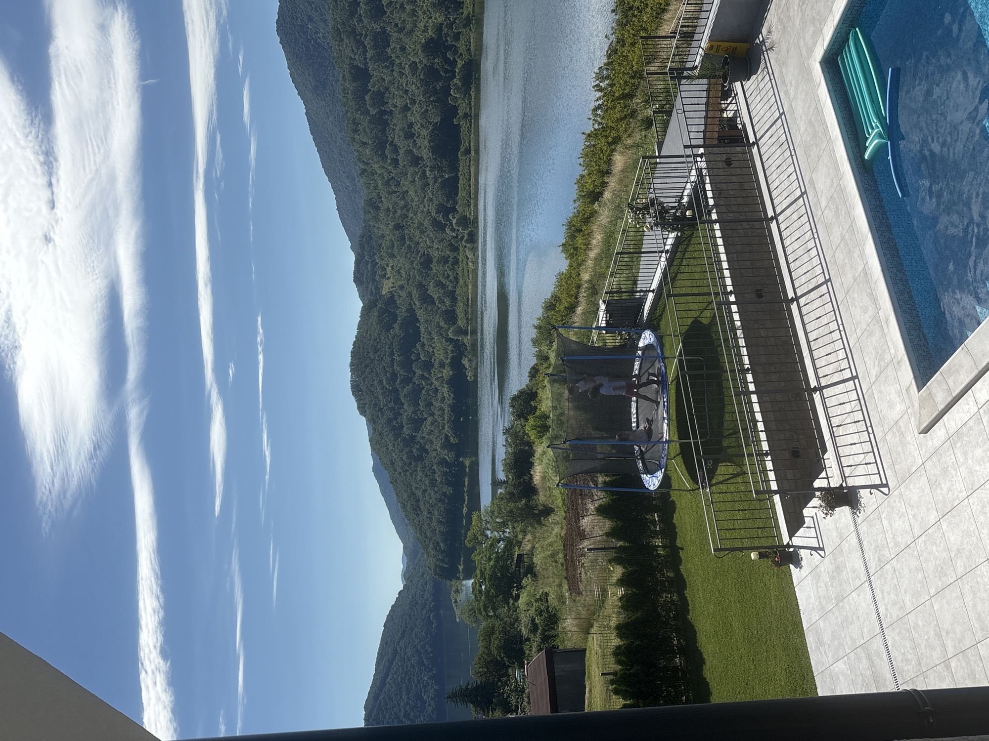 This is an eye-level, outdoor, daytime, medium shot of a residential property with a lake and mountains in the background. The scene is divided into foreground, midground, and background elements. In the foreground, a tiled patio leads to a swimming pool containing a blue inflatable pool float. A black metal railing encloses the patio area. In the midground, a green lawn is visible, with a trampoline in the center occupied by two children. A small shed is situated to the left of the trampoline, with a fence running behind the lawn. Beyond the lawn, the ground slopes down to a body of water, bordered by lush green trees. In the background, rolling, tree-covered mountains rise from the edge of the water, under a bright blue sky with wispy white clouds.