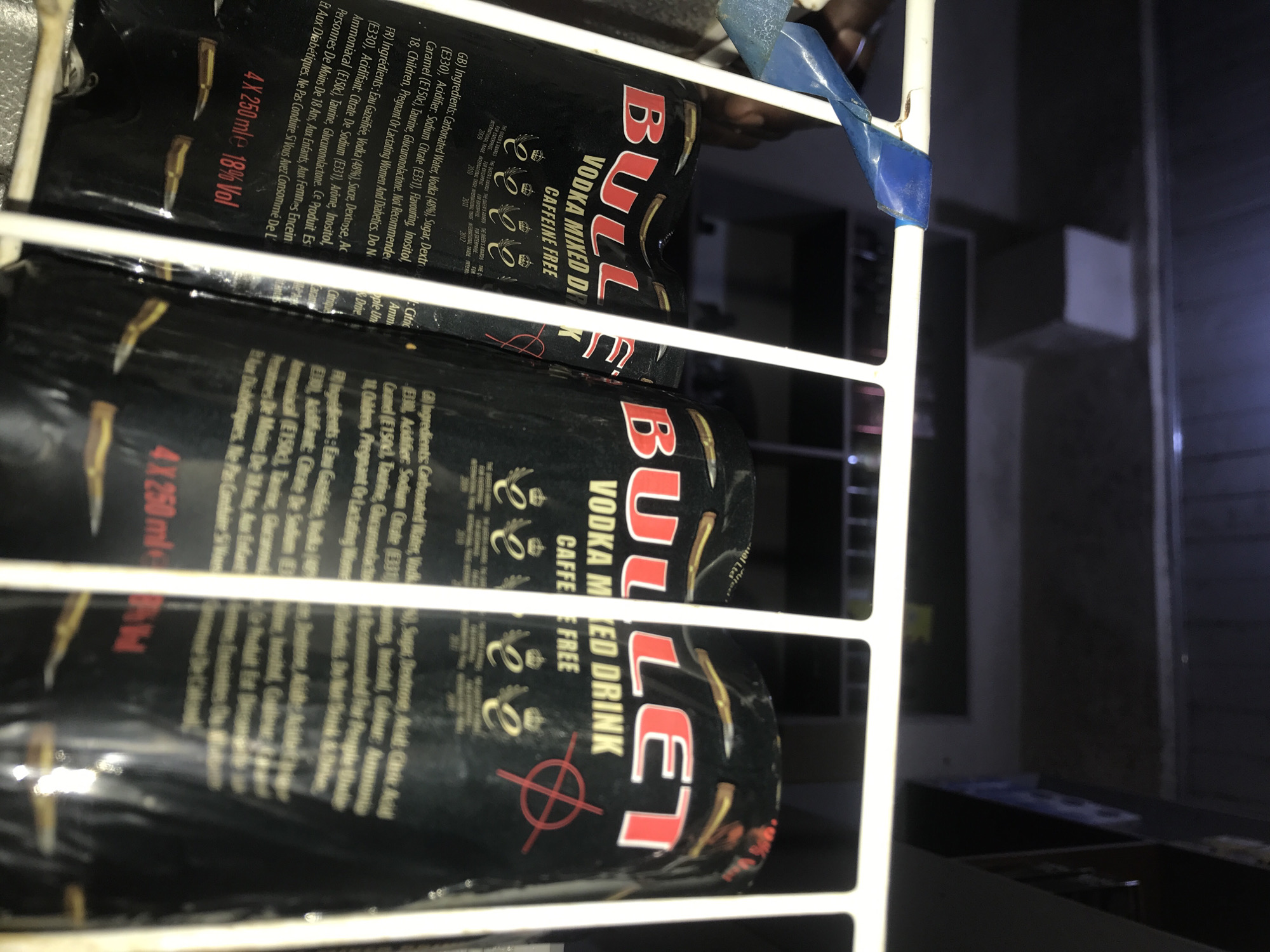 The image shows a close-up, low-angle shot of three cans of "Bull" vodka mixed drink with caffeine-free packaging. The cans are black with red and gold lettering and graphics, including a stylized bull logo and bullet designs. The cans are arranged in a row and appear to be inside a refrigerator or display case, indicated by the white metal bars in the foreground. The lighting is dim, suggesting an indoor environment, possibly a store or bar. There are no discernible people or specific location details visible to confirm it being in New Stadium, Nigeria. The text on the cans includes ingredient lists in English and French, as well as warnings. The overall scene is focused on the product packaging.