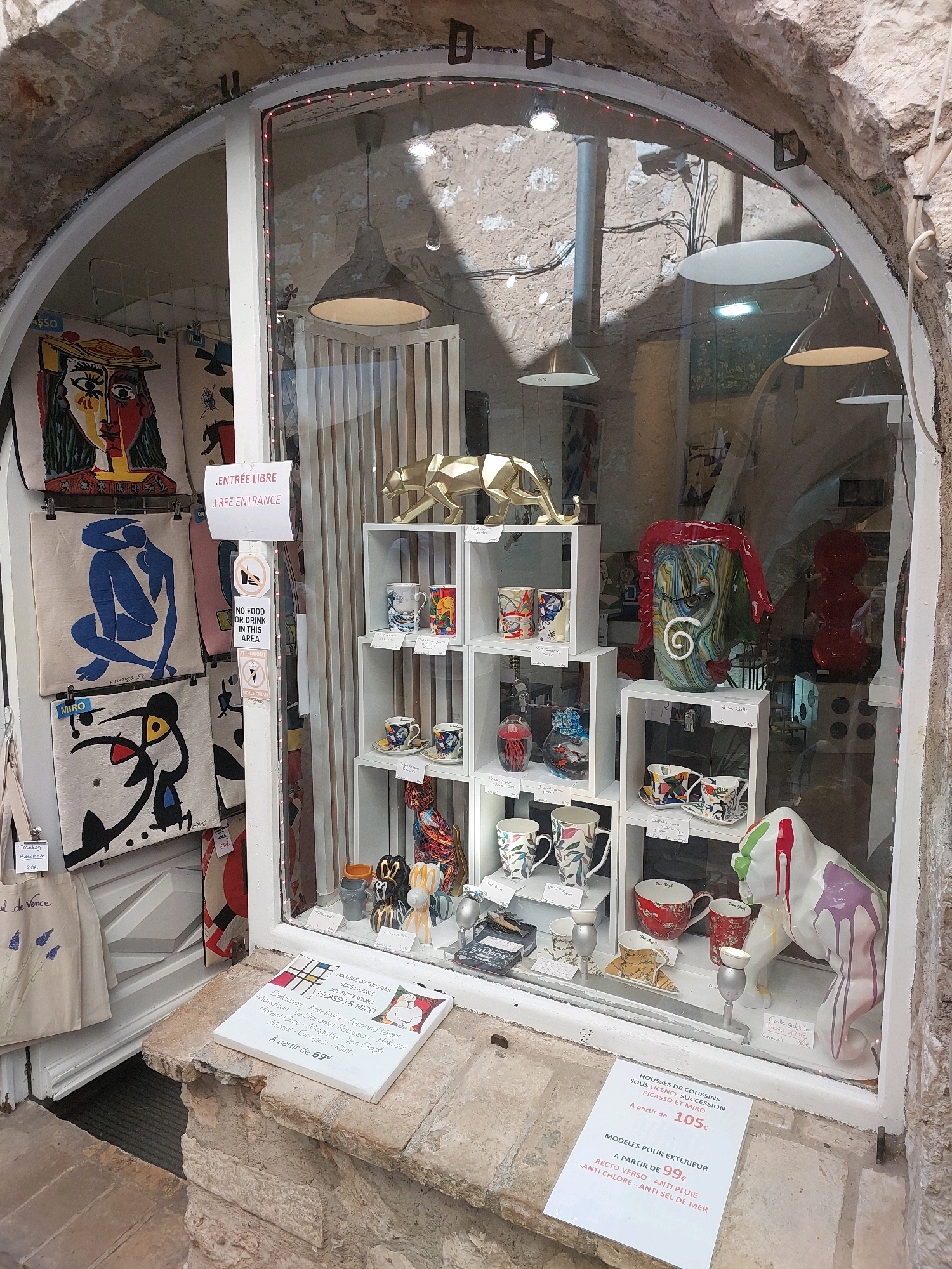 The image shows a storefront display in Saint-Paul-de-Vence, France. The display features a variety of art and decorative items, including cushions with reproductions of artworks by Picasso and Miro, as well as ceramic mugs, vases, and sculptures. A white sign on the right side of the display advertises "MODÈLES POUR EXTÉRIEUR" (Outdoor models) with prices starting from 99€. The storefront has an arched entrance with rough stone walls, and the window display is illuminated by interior lighting. There are no people visible in the scene.