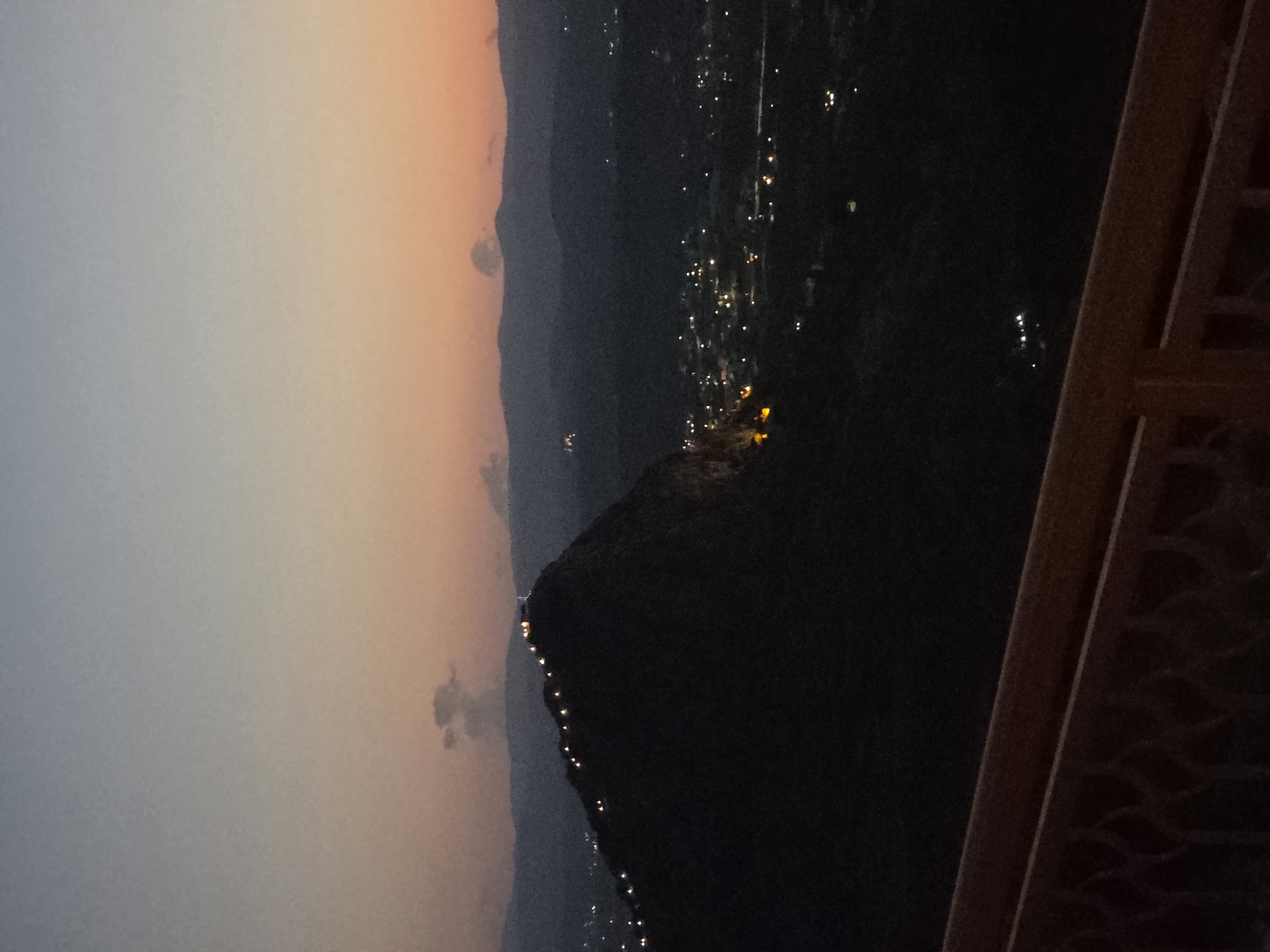 The image captures a twilight view from an elevated position overlooking a landscape that includes a prominent, dark-hued mountain or hill. The sky transitions from a pale blue at the top to a soft orange and pink hue near the horizon. Numerous small, glowing lights are visible along the crest of the mountain, suggesting a pathway or structures. Further in the distance, the lights of a settlement or town are scattered across a valley. The foreground features a wooden railing, indicating the vantage point is likely a balcony or observation deck. The overall scene depicts a serene evening setting with artificial lights illuminating the darkness.