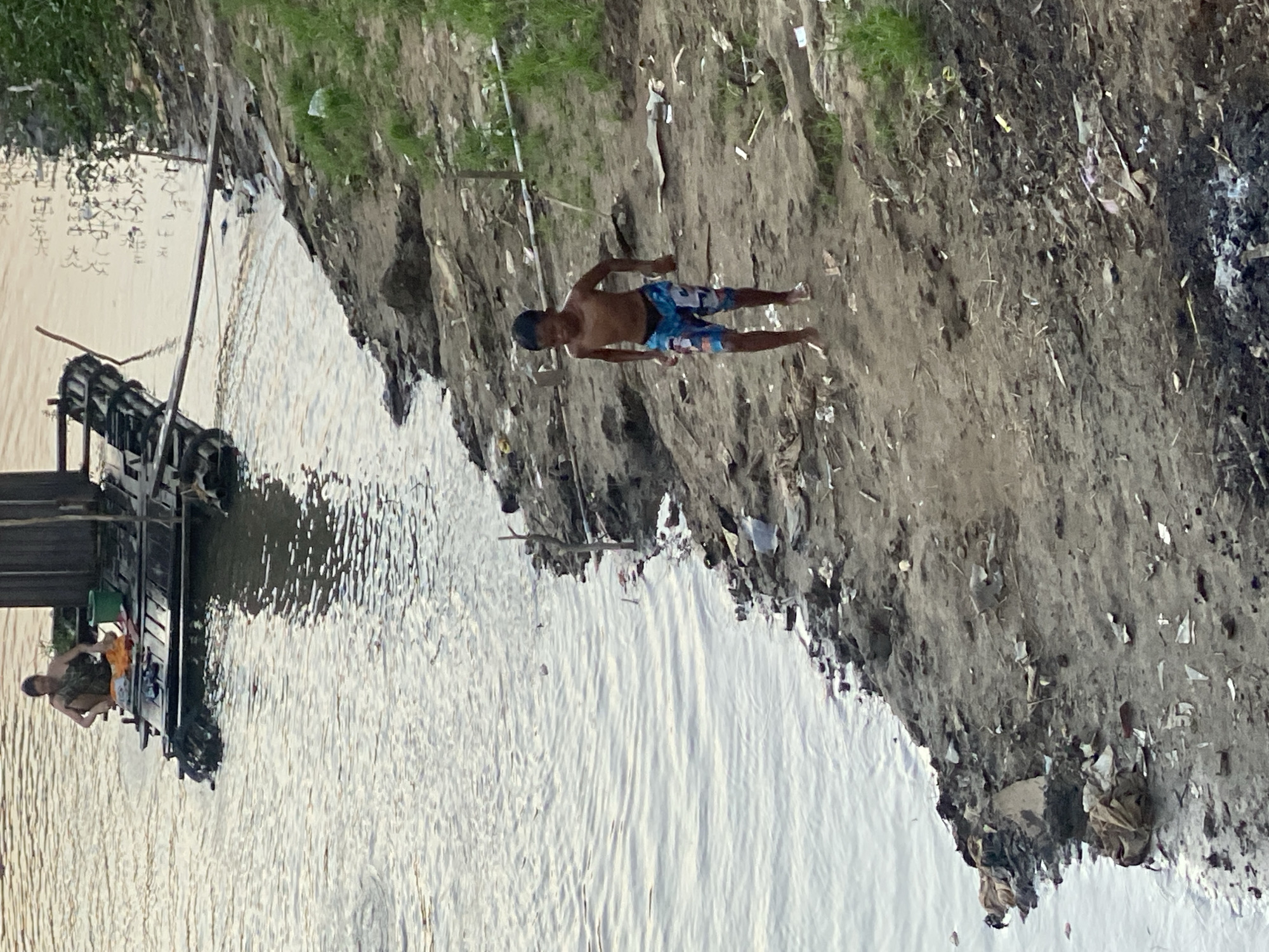 An elevated, eye-level shot shows a young boy running on a muddy, trash-strewn riverbank in Tunggul Irang Ulu, Indonesia. The boy, who is shirtless and wearing patterned swim trunks, is moving from right to left across the frame, his arms slightly bent and his gaze directed forward. The riverbank is uneven and littered with various debris, including plastic, fabric, and what appears to be burnt material in the foreground. In the background, on the river, a raft is visible. A person is seated on the raft, facing away from the camera, appearing to be washing clothes or engaging in a similar activity. A green bucket is also present on the raft. The water is calm, reflecting the muted light of what seems to be either early morning or late afternoon. The environment suggests a scene of poverty or informal settlement along the river.