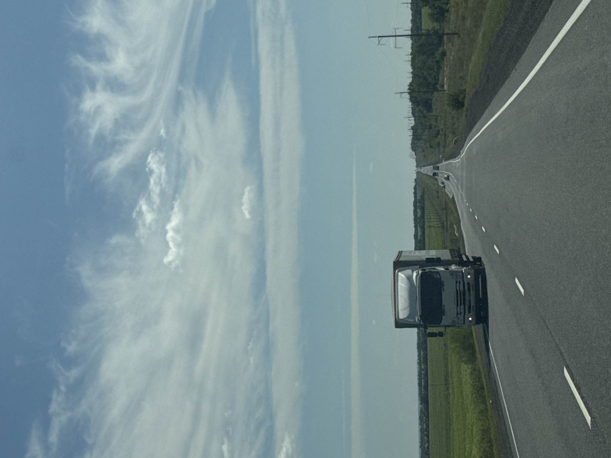The image shows a two-lane asphalt road stretching into the distance in Burdygino, Russia. The sky is blue with scattered white clouds. On the left side of the road, there is a green field. On the right side, there are power lines and trees. In the foreground, a white truck with a trailer is traveling on the road. Further down the road, other vehicles are visible. The overall scene depicts a typical rural highway in Russia on a clear day.