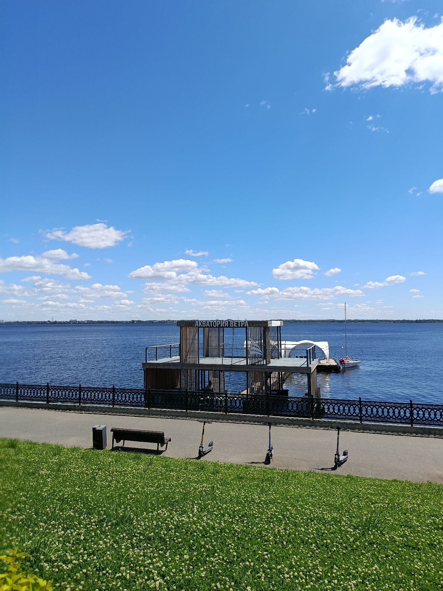 This image depicts a waterfront scene in Saratov, Russia. In the foreground, a lush green lawn covered in small white flowers slopes down towards a paved walkway. Along the walkway, there is a dark trash receptacle, a wooden bench, and two electric scooters parked in a row. A decorative black metal fence separates the walkway from the body of water.
Dominating the middle ground is a multi-level wooden structure situated on the water, appearing to be a pier or a cafe. The structure has a modern design with open-air sections and a white canopy. A small sailboat with a red-clad person on board is docked near the structure. The word "АКВАТОРИЯ" (AkVATORIYa), meaning "Aquatoria," is visible on the side of the structure.
The background features a wide expanse of blue water, with a distant shoreline visible on the horizon, lined with trees and buildings. The sky above is a clear, bright blue, dotted with scattered white cumulus clouds. The overall impression is a sunny day in a public park or promenade area.