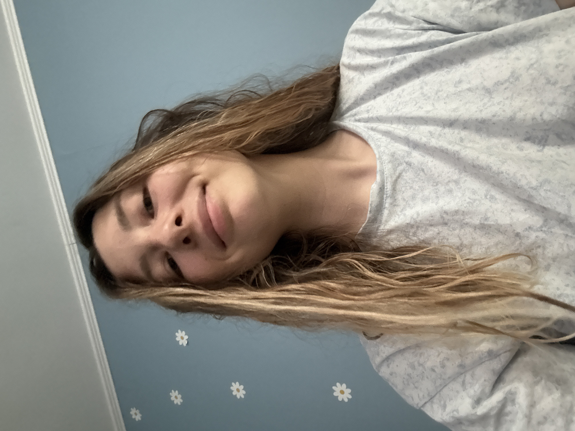 A medium shot captures a young woman from the chest up, looking slightly upwards towards the camera. She has long, light brown, wavy hair that falls around her shoulders. Her facial expression is a gentle smile, with her head tilted slightly to her left. She is wearing a light gray t-shirt with a subtle, mottled pattern. The background is a pale blue wall adorned with white daisy-shaped stickers. A white decorative trim runs along the top of the wall where it meets the ceiling. The lighting suggests an indoor environment, likely during the daytime. The image does not contain any information that would allow for identification of a specific location in Moscow, Russia.