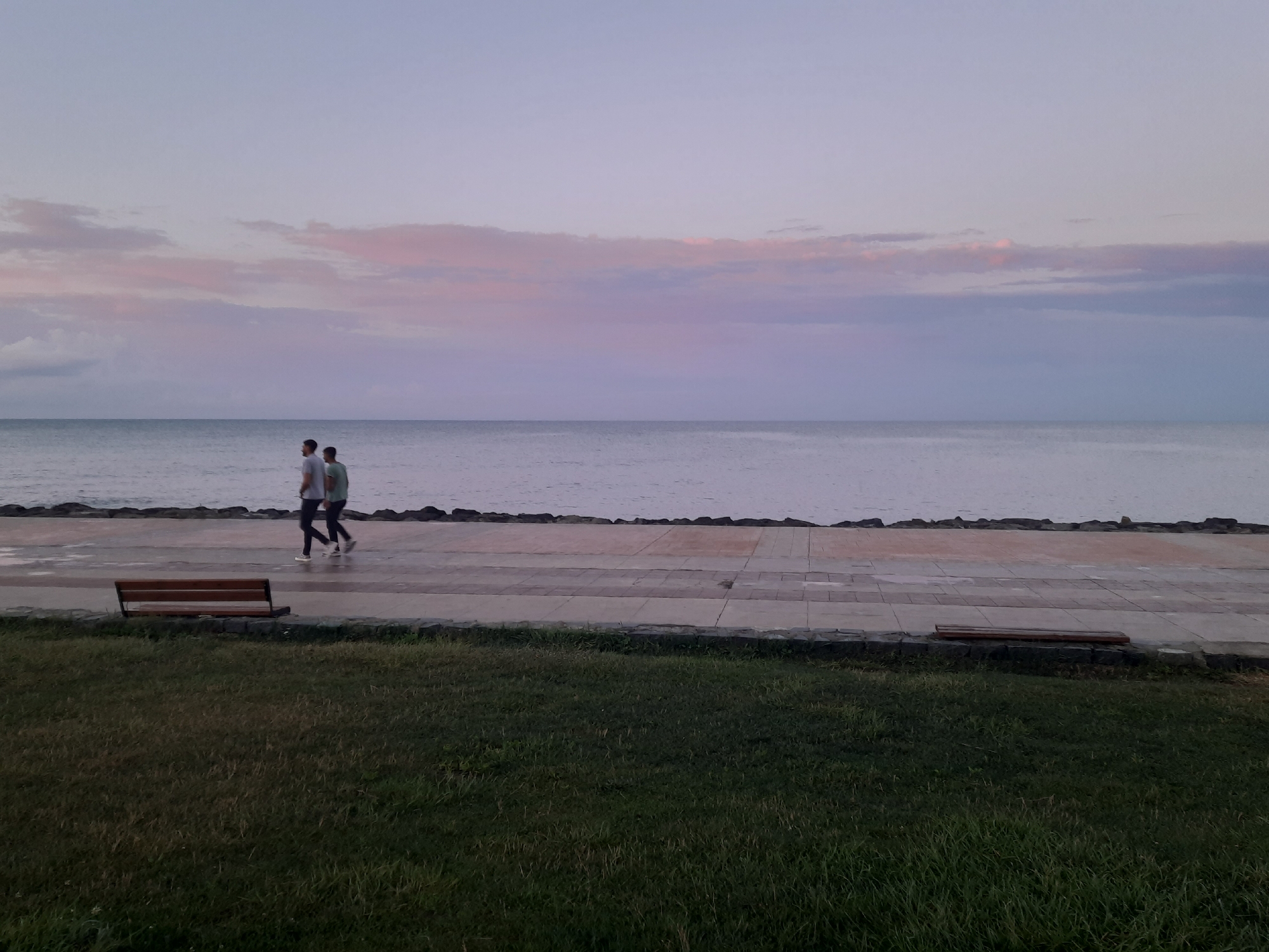 At dusk, two individuals walk side-by-side along a paved promenade bordering the sea in Atakum, Turkey. The sky is a soft gradient of pale lavender and pink, with wispy clouds. The sea is calm and reflects the muted sky. A low stone wall separates the promenade from a grassy area. A wooden bench is visible on the promenade, and another bench is to the right of the frame. The overall scene evokes a peaceful evening ambiance.