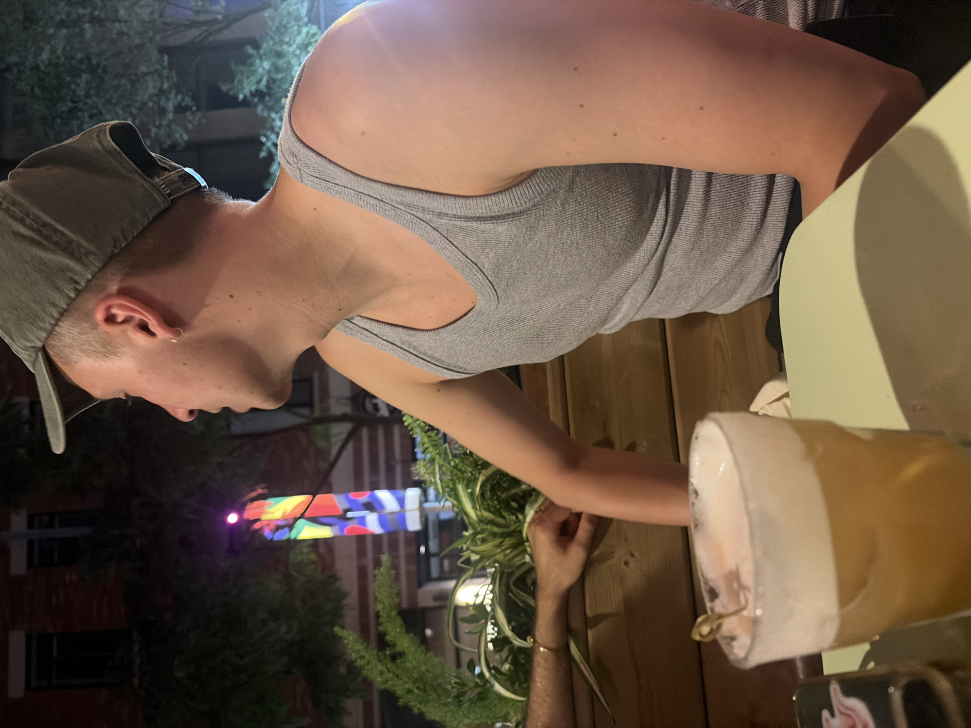 The image shows a young man wearing a grey tank top and a green cap, looking to his right. He has a small hoop earring in his left ear. He is sitting at a table with a drink in a glass that has a foamy top. In the background, there is a building with brick facade and windows, a tree, and a colorful light installation. The scene appears to be an outdoor seating area of a restaurant or bar at night. The reflection on the glass suggests it's a warm evening. This specific location is a restaurant on Saint-Laurent Boulevard in Montreal, Canada, known for its vibrant nightlife and artistic decorations.
