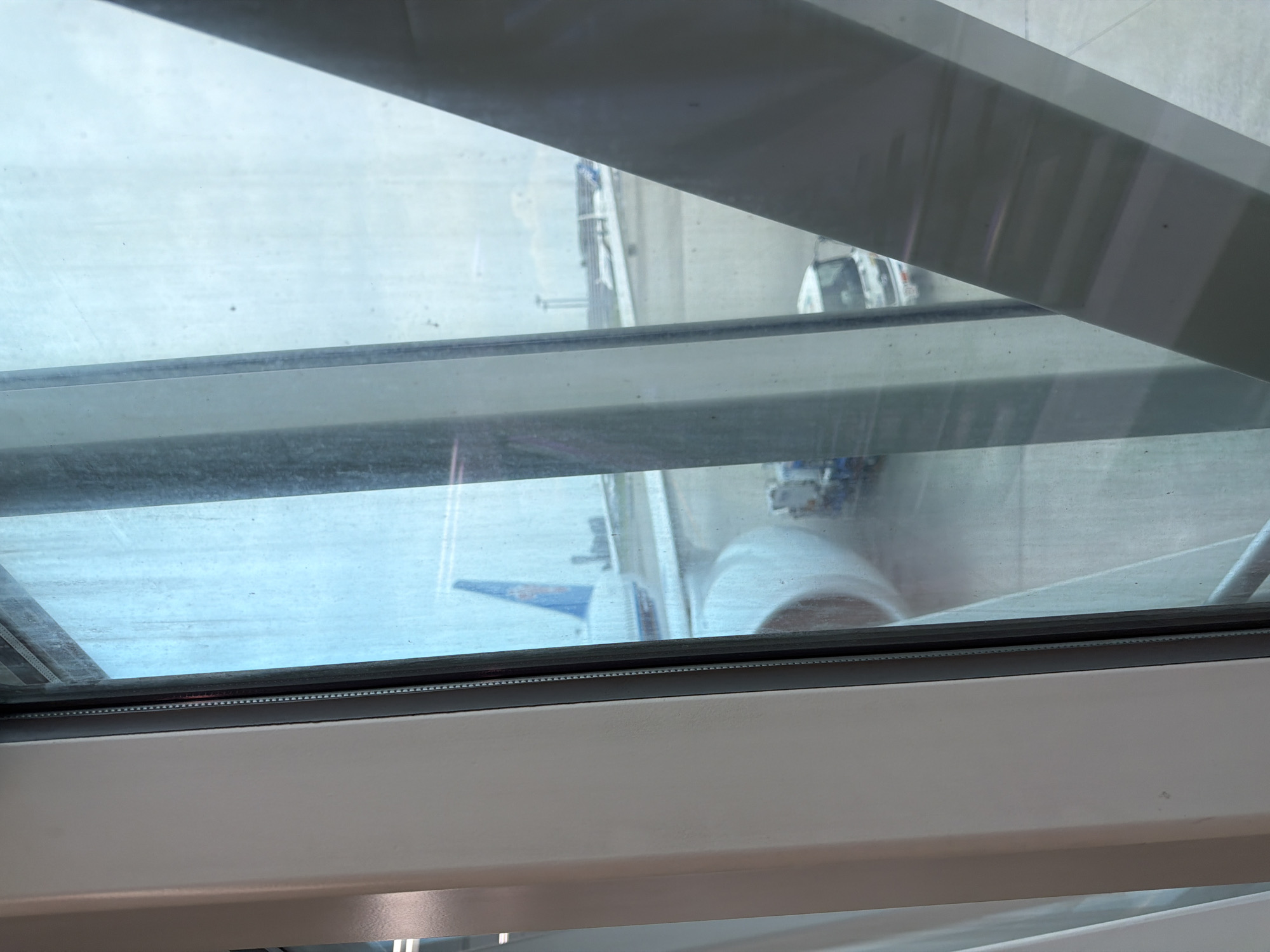 A view from inside an airport jet bridge shows an airplane parked on the tarmac. The tail of the airplane, featuring a blue and orange logo, is visible. A service vehicle is parked near the airplane. The image is taken from inside a jet bridge, looking out through the window at the airport scene. The location is Ota, Japan, likely at Haneda Airport.