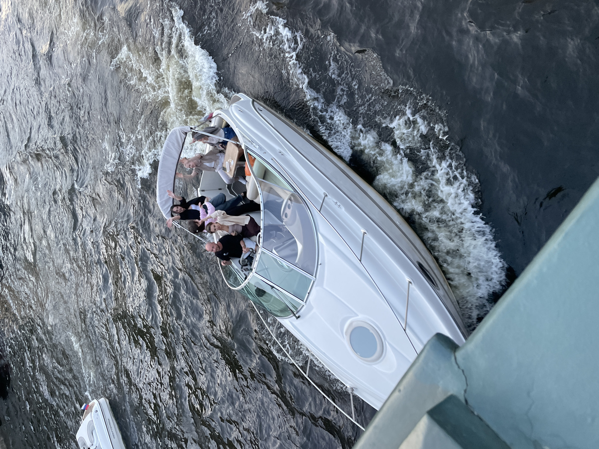 A white motor yacht, carrying seven individuals (one man and six women), navigates a dark, choppy waterway in Saint Petersburg, Russia. The man is positioned at the helm, wearing a dark t-shirt. Several women, dressed in casual attire, are seated in the stern, with some waving towards the camera. The yacht features a white hull, a grey bimini top, and a glass windshield, generating a significant white wake behind it. In the upper left background, another smaller white boat is visible, also moving, with a Russian flag affixed to its stern. The scene is viewed from an elevated perspective, likely a bridge, with a light blue-green metal railing prominent in the foreground, displaying a visible crack on its upper horizontal surface. The water exhibits distinct ripples and waves from boat movement.