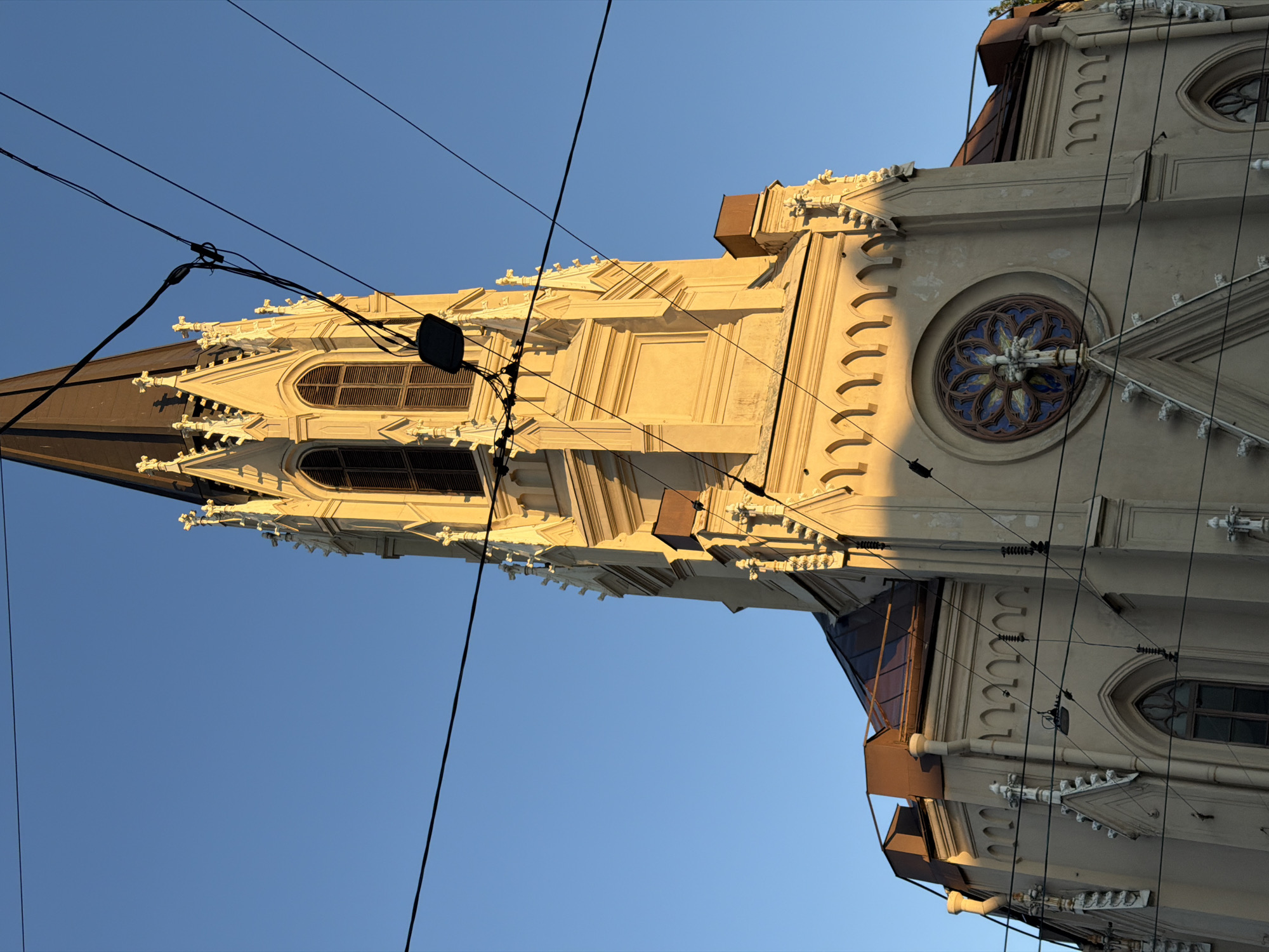 The image depicts a low-angle view of the upper sections of the Catholic Church of St. Catherine, situated on Nevsky Prospekt in Saint Petersburg, Russia. The church, constructed in a Neo-Gothic architectural style, features a prominent tall, slender spire with a dark, pointed roof that dominates the upper portion of the frame. The building's facade is primarily light cream or beige, illuminated by warm, direct sunlight, creating distinct shadows that highlight its intricate details.
The spire includes several pointed turrets and decorative finials along its edges. Below the spire, multiple tall, arched windows fitted with dark louvers are visible. A large circular rose window, centrally positioned on the lower visible facade, displays ornate blue, brown, and possibly white stained glass with a cross design at its core. Further architectural elements include numerous pointed arches, small decorative columns, and cornices. Dark, sloped roof sections, possibly metal, are visible on lower parts of the structure, with a few rectangular, brown ventilation or skylight elements present.
The background is a clear, vibrant blue sky. Several black utility wires, likely tram or electrical lines, crisscross diagonally and horizontally across the foreground, partially obscuring the church. Some wires exhibit insulators and connectors, and a small black rectangular device is suspended from one of these wire clusters. No individuals are visible in the scene.
