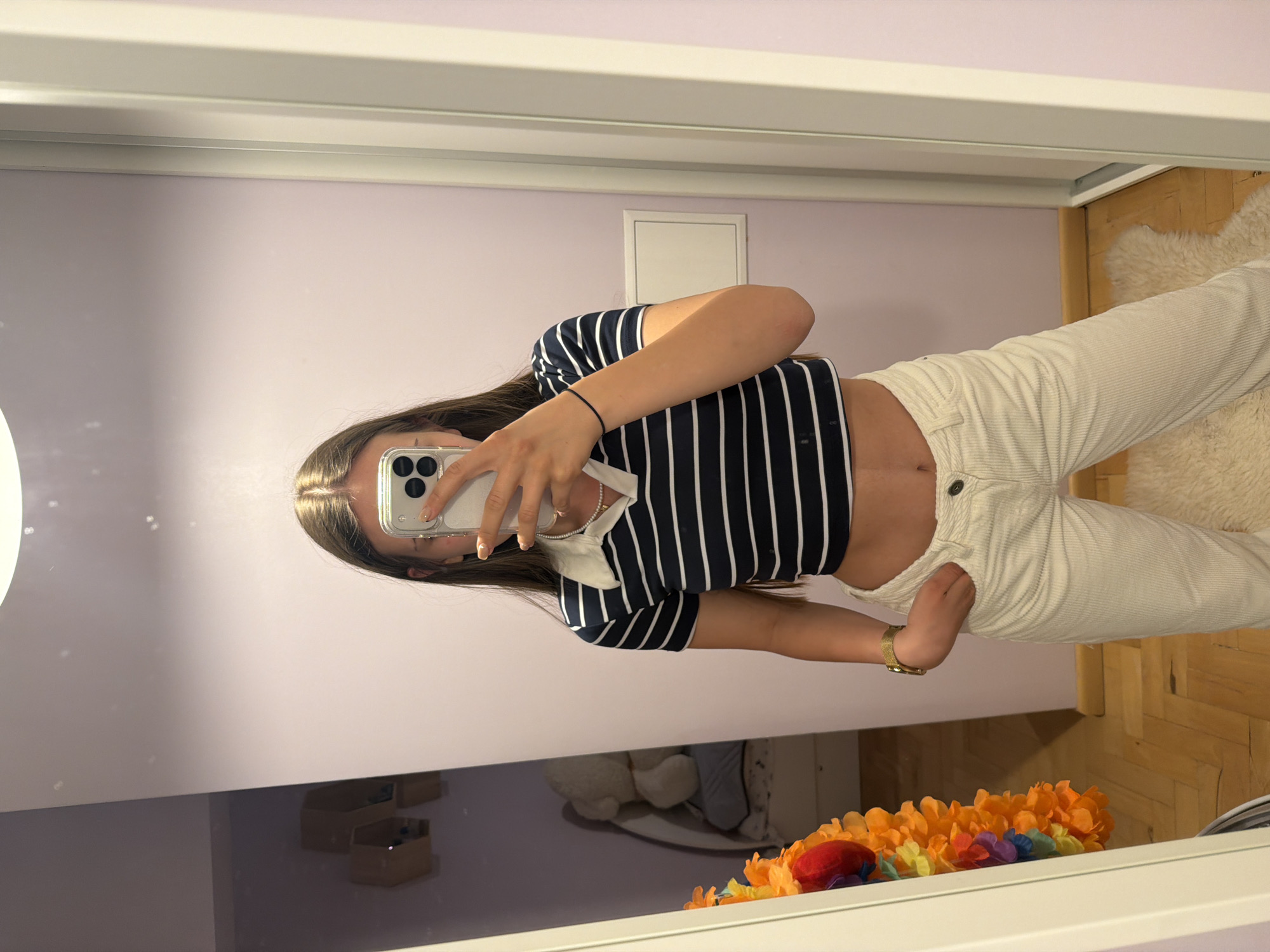 The image shows a young woman taking a selfie in a mirror. The woman is wearing a navy and white striped shirt and cream-colored pants. She is holding a smartphone up to her face, obscuring her features. Her hair is long and brown. The background of the image includes a light purple wall, a white door or panel, a wooden floor, a white rug, and a garland of orange and red flowers. A reflection in the mirror also reveals some storage boxes and a plush toy. The location is not discernible as Buhovo, Bulgaria.