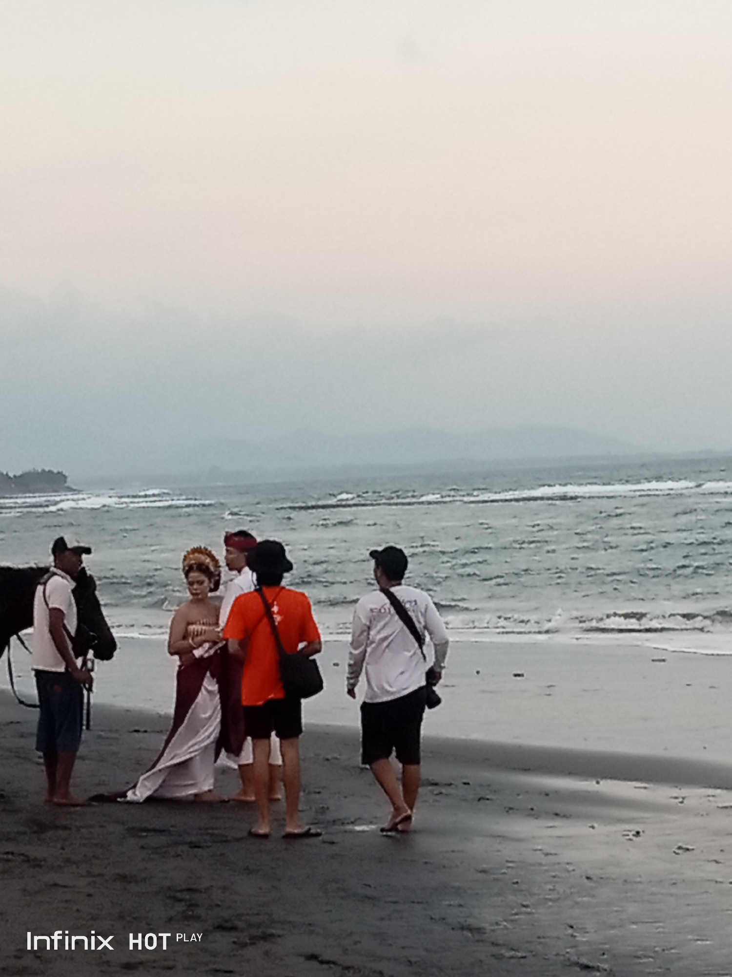 In Blahbatuh, Indonesia, on a dark sand beach, a group of people is gathered near the ocean. A man in a white t-shirt and blue shorts stands to the left, next to a black horse. Beside him, a woman in traditional attire with a golden headdress and a dark sarong is being attended to by another person in a red outfit. To the right, a man in a white long-sleeved shirt and black shorts walks away from the camera. The ocean waves are visible in the background, with distant mountains obscured by haze. The sky is overcast, suggesting either early morning or late afternoon.