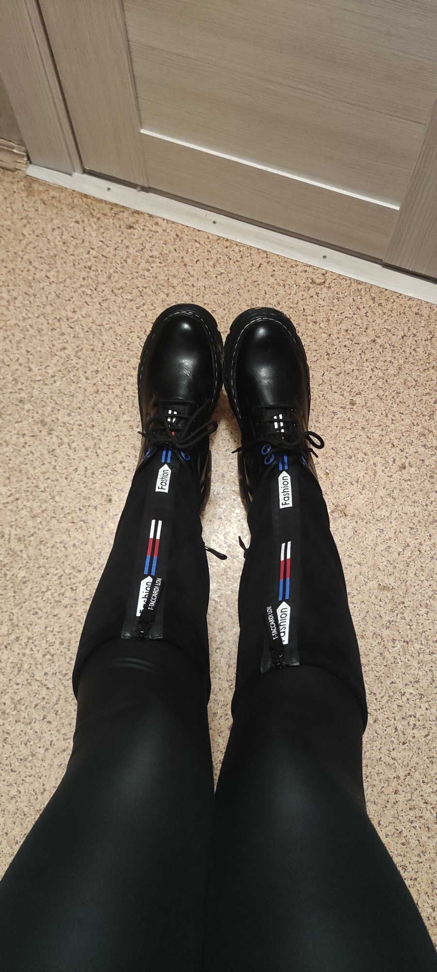 The image shows a person standing indoors, wearing black leggings and black leather boots with white "Fashion" labeling and zippers on the outer sides of the lower legs. The boots are laced with black and have blue accents. The flooring is a tan speckled material, likely linoleum or a similar synthetic. A door with a light wood grain finish is visible in the background. The location is likely a residence in Chaikovsky, Russia, based on the origin of the image.