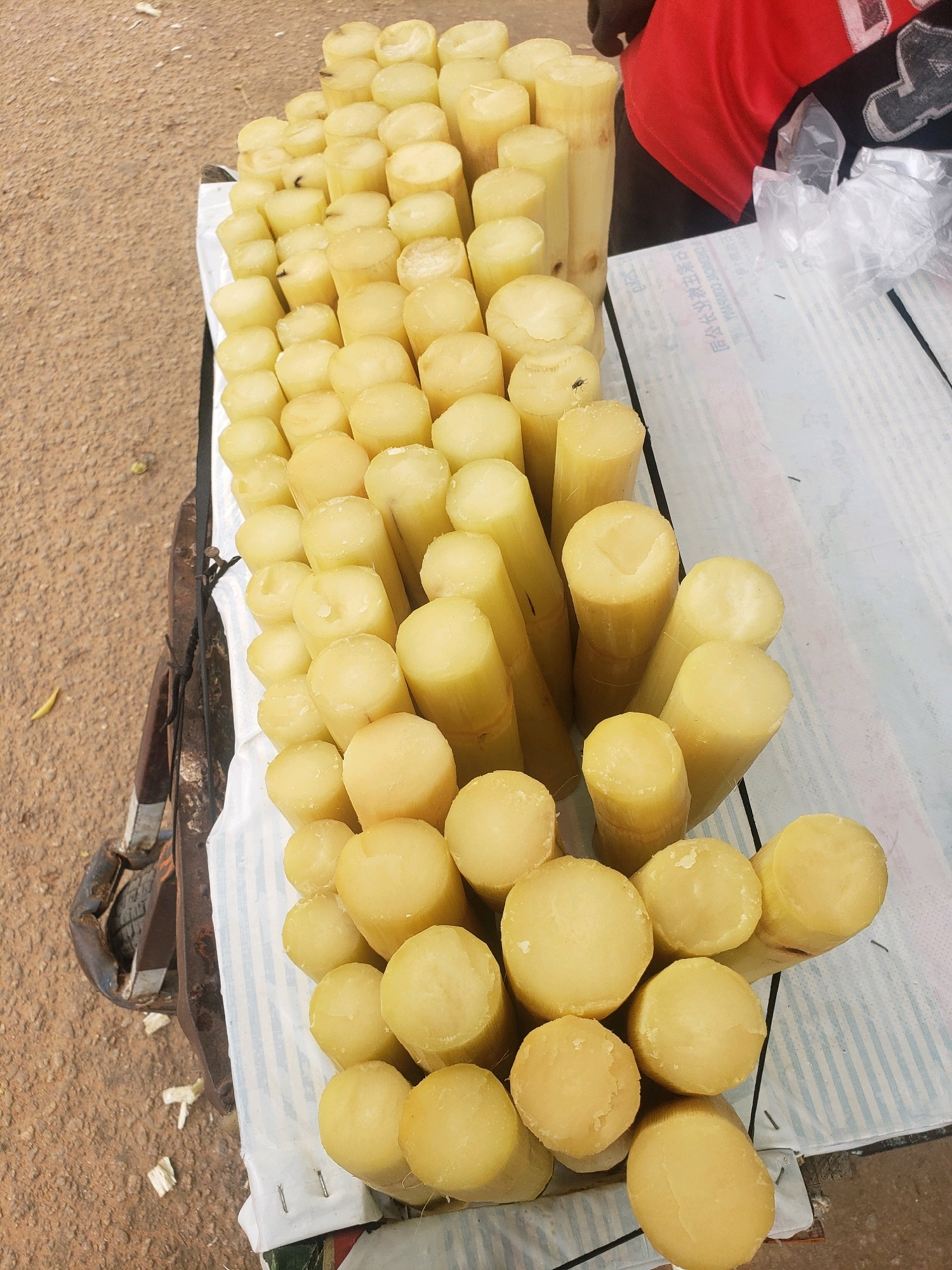 In Narayi, Nigeria, a vendor displays a pile of sugarcane stalks. The sugarcane is cut into manageable segments, varying in thickness and length, and is arranged on a white cloth laid over a wooden surface. The stalks are a pale yellow color, with some showing rough, fibrous ends. A small fly is visible on one of the sugarcane pieces. In the background, a person wearing a red shirt is partially visible, and transparent plastic bags are also present. The ground is a sandy, reddish-brown color. A part of a cart or stand is visible on the left side of the image, with a tire and metal frame.