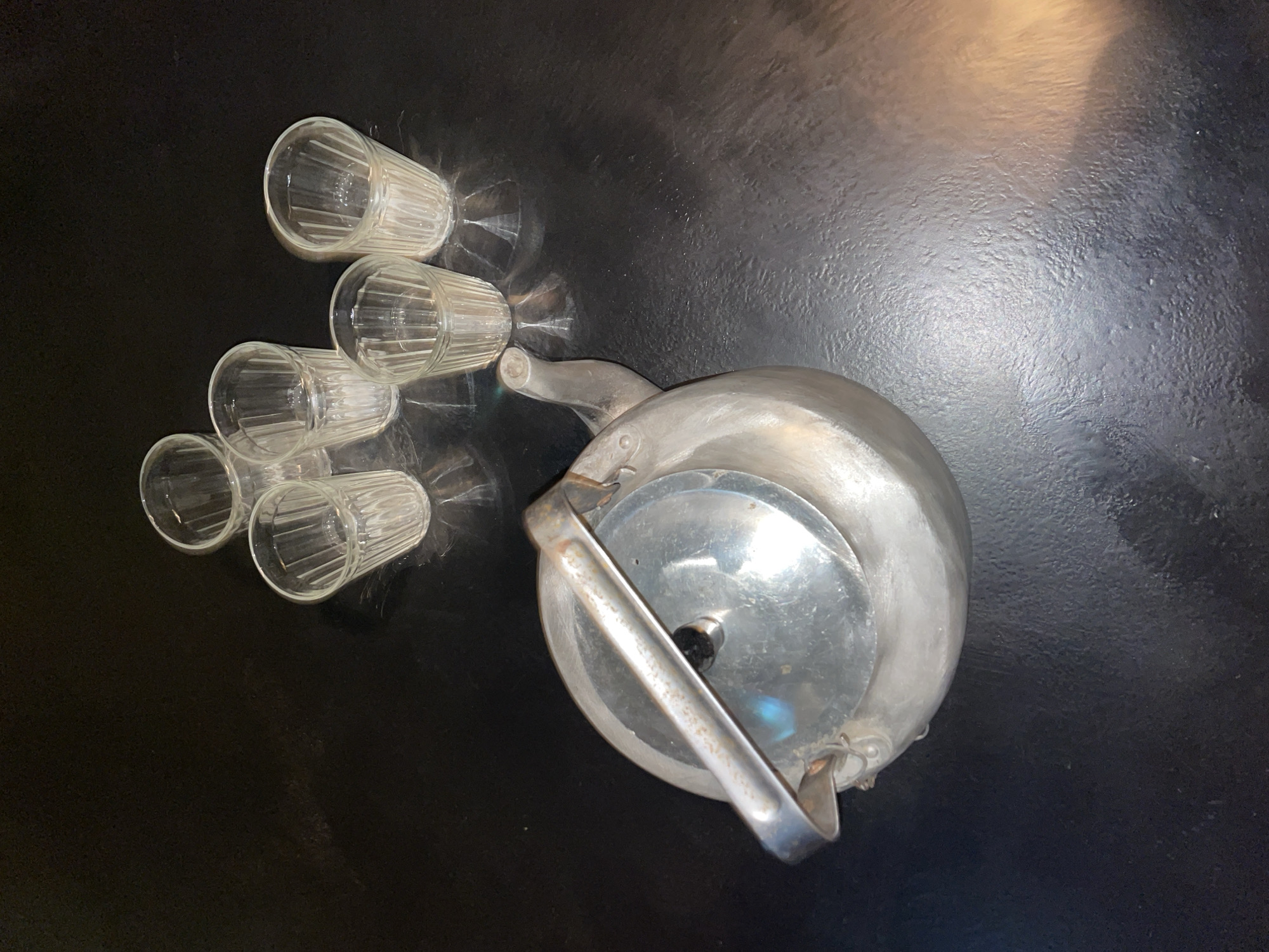 The image presents a high-angle shot of several glass cups and a teapot on a dark, possibly black, surface. There are five clear, ribbed glass cups arranged in a cluster on the left side. To the right of the glasses sits a metal teapot with a hinged lid and a handle. The lighting creates reflections on the metallic surface of the teapot and within the glasses, adding highlights and shadows to the scene. There are no people visible in the image. The scene appears indoors. There is no specific information in the image to identify its location as Moscow, Russia.