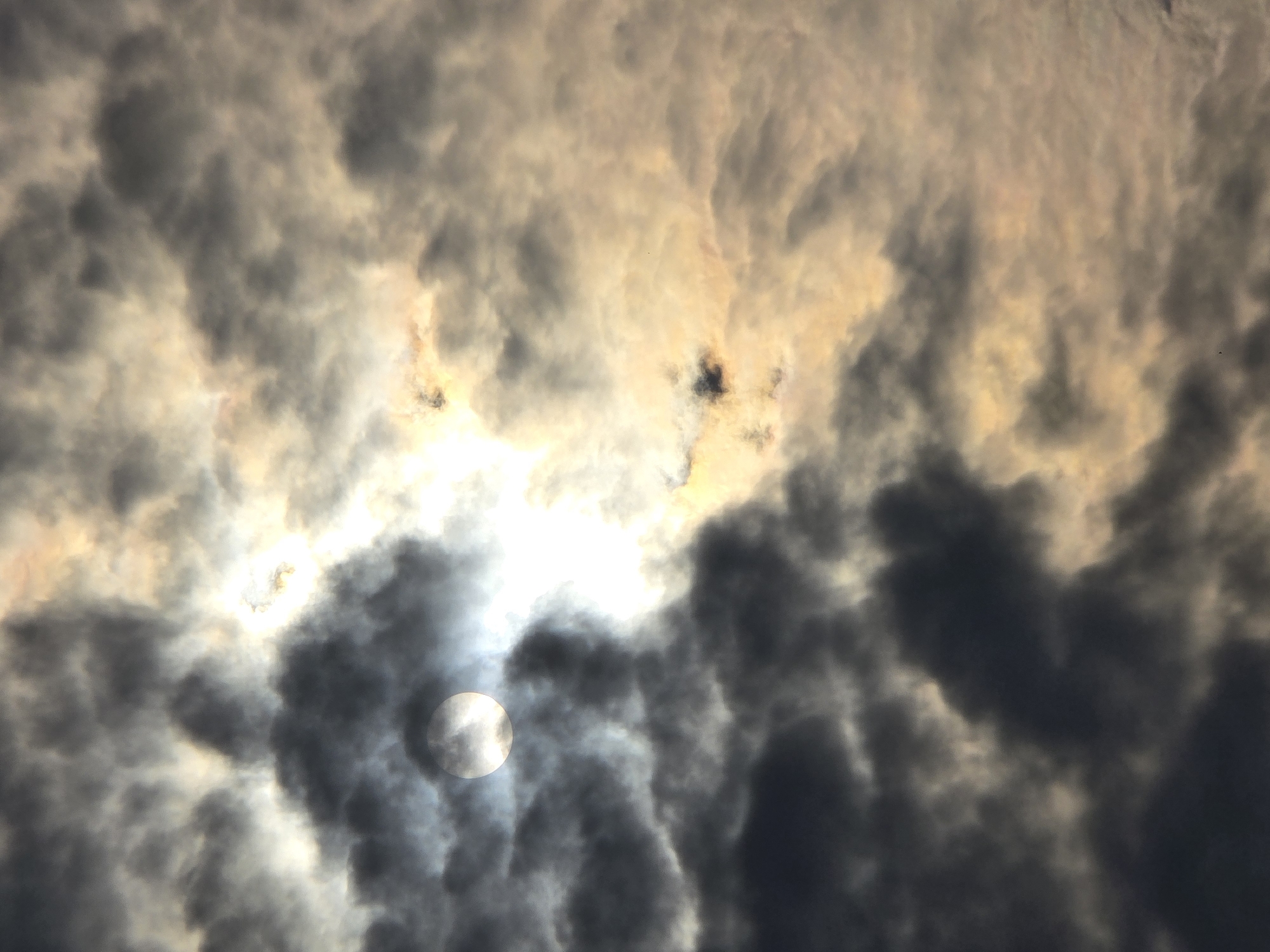 The image captures a dramatic sky filled with a mixture of dark, heavy clouds and brighter, sunlit areas. The sun is partially obscured by a dark cloud, creating a halo effect and casting a bright, diffused light. The clouds are textured and varied, with some appearing soft and wispy, while others are dense and almost menacing. The lighting suggests either sunrise or sunset, with warm, golden tones mingling with cooler grays and blacks. The overall impression is one of dynamic weather conditions, perhaps a storm passing or approaching. The location in Ермолаево, Russia, is not visually depicted within the image itself; it is a contextual detail provided. There are no people, identifiable structures, or specific objects visible in the frame, only the natural elements of the sky.