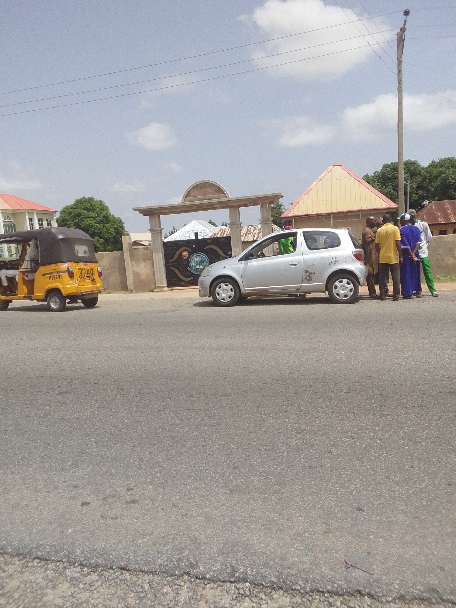 In Jalingo, Nigeria, a yellow auto-rickshaw with the license plate PT324R is parked on the side of a road. In front of it, a silver car is stopped, and a group of five men are gathered around it, appearing to be in conversation. The men are dressed in various traditional African attire and western casual clothing. Behind them, an arched gateway with an ornate gate leads to a compound. The gateway is flanked by stone pillars and a large, decorated gate with a crest. Adjacent to the gateway is a building with a distinct yellow, triangular roof. The scene is set under a bright, cloudy sky with utility poles and wires visible in the background. The road is paved, and the surrounding environment appears to be a mix of urban and suburban elements with trees and other buildings in the distance.
