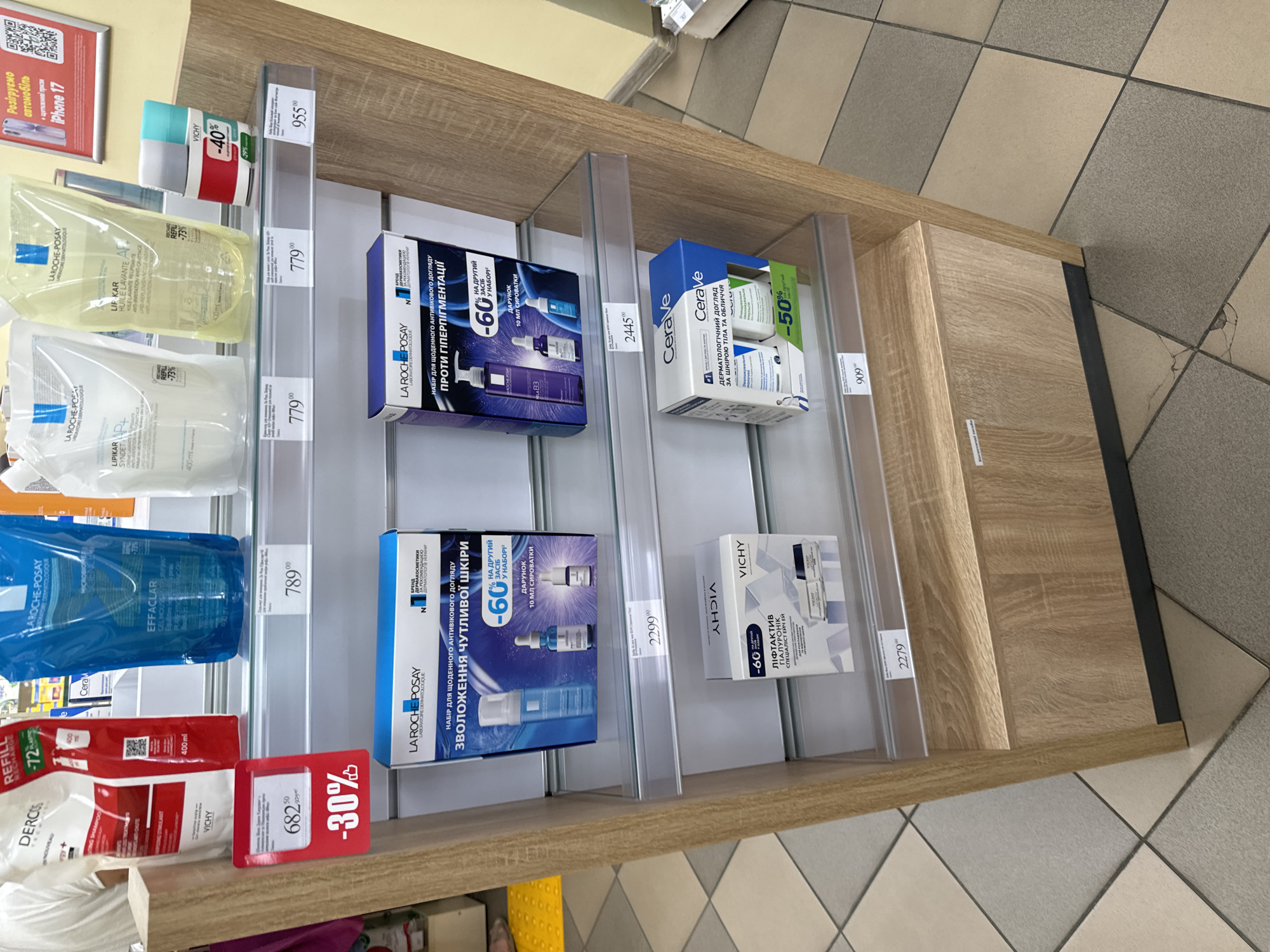 This is a photograph of a retail display shelf in a store, likely a pharmacy or beauty store, in Sarny, Ukraine. The shelf unit is made of wood-grain laminate with clear glass shelves. On the shelves are various skincare products, primarily from the brands La Roche-Posay and Vichy, as well as a Cerave product.
The top shelf displays several large pouches of liquid products, including La Roche-Posay Effaclar and Lipikar. There are also smaller Vichy bottles with discount stickers showing "-40%" and "-20%". A price tag of "955" is visible on this shelf.
The second shelf holds a La Roche-Posay set for sensitive skin, with a price tag of "2299".
The third shelf features a La Roche-Posay set for hyperpigmentation, with a price tag of "2445".
The bottom shelf displays a Vichy set for lifting and a Cerave set, with price tags of "2279" and "909" respectively. A red discount tag showing "-30%" and a price of "682" is attached to the wooden support structure on the left side of the display.
The floor is tiled with a pattern of beige and grey squares. The overall lighting is bright, suggesting an indoor commercial environment. No people are visible in the frame.