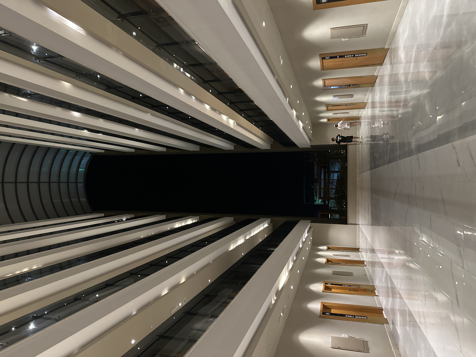 The image depicts a modern, multi-story atrium or hallway with a high ceiling, likely at night. The architecture features repeating white horizontal bands that create a sense of depth and perspective, leading towards a dark, expansive area at the top. The floors are highly reflective, showing a marble-like pattern.
On the left and right sides of the hallway, there are multiple doors, each with a wooden finish and a label indicating a room number. The doors are evenly spaced, and some have small rectangular panels mounted on the wall next to them. Lighting fixtures are embedded in the ceiling and along the sides of the upper levels, casting a warm glow.
In the lower right portion of the image, two individuals are walking away from the camera. One is wearing a black t-shirt and dark shorts, and the other is wearing a white t-shirt and light-colored shorts. Their reflections are visible on the glossy floor.
In the far background, beyond the immediate hallway, there appears to be a glimpse of an outdoor area with some structures and lights, suggesting the location is part of a larger building complex or a hotel.
While the image itself does not contain any explicit clues to its location in Manavgat, Turkey, the architectural style and interior design are consistent with modern hotel or resort facilities commonly found in tourist destinations like Manavgat.