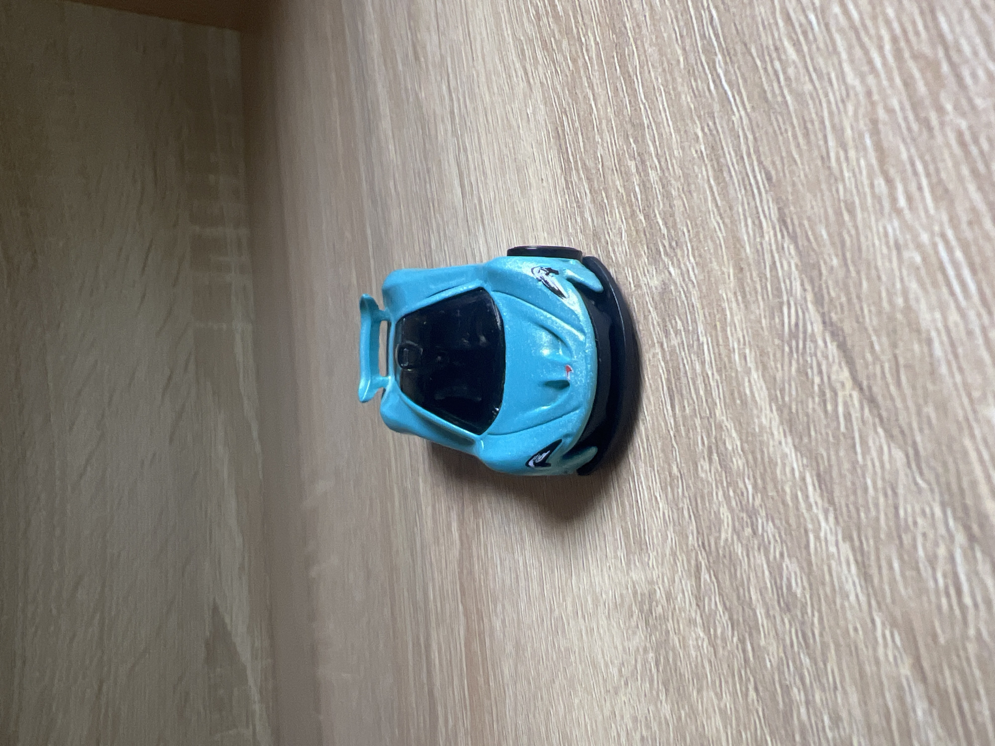 A blue toy car sits on a light brown wooden surface. The car is facing the camera and is slightly angled to the left. The surface appears to be a table or shelf, with a wooden grain pattern visible. In the background, there is a lighter brown surface, possibly another shelf or wall. There is no indication of the city of Minsk, Belarus, or any other specific location. The scene is static and shows no action or interaction.