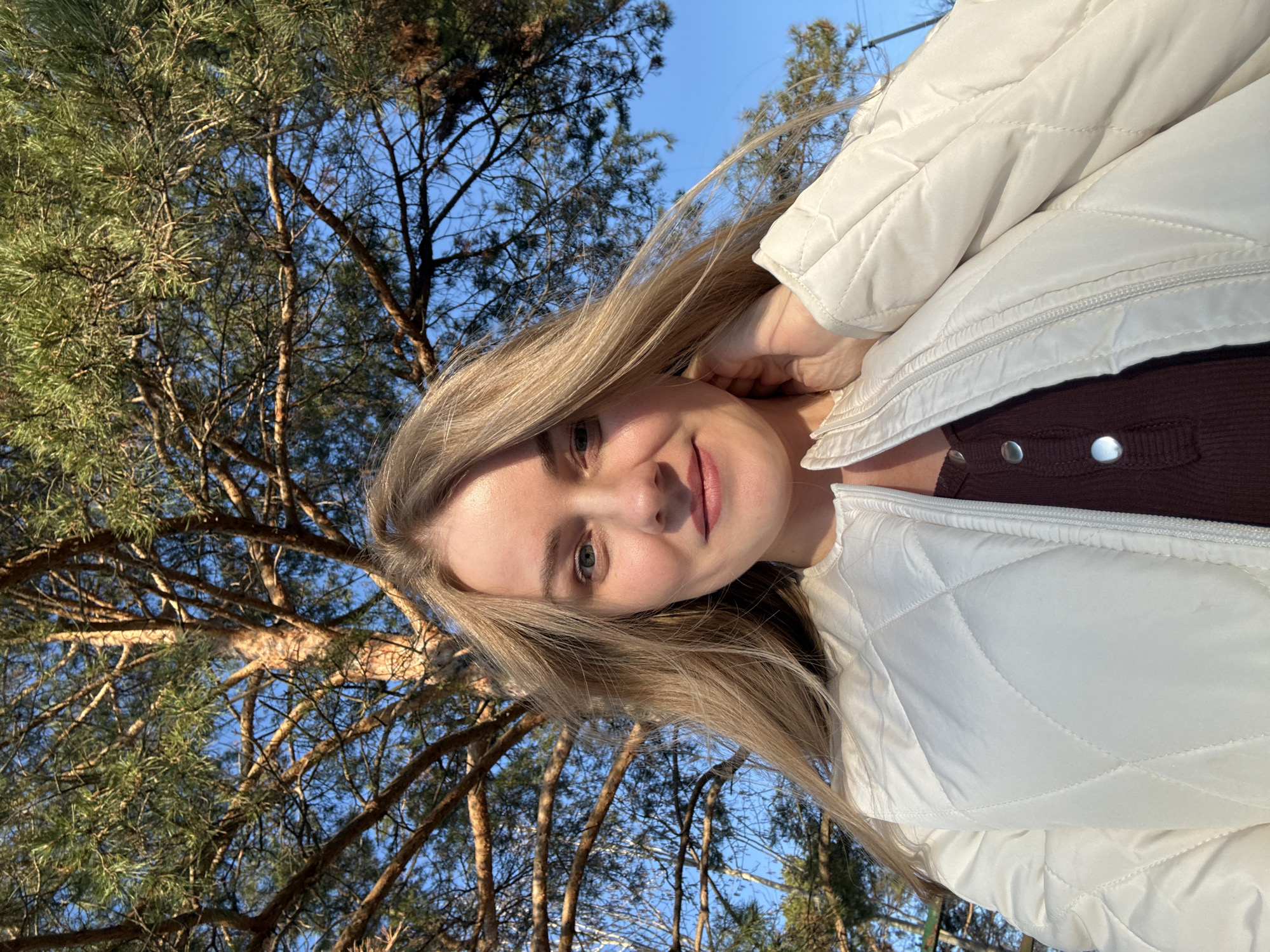 A woman poses for a selfie outdoors in Chaikovsky, Russia. The background features trees and a blue sky. She wears a white quilted jacket over a brown top. Her blonde hair is shoulder-length.