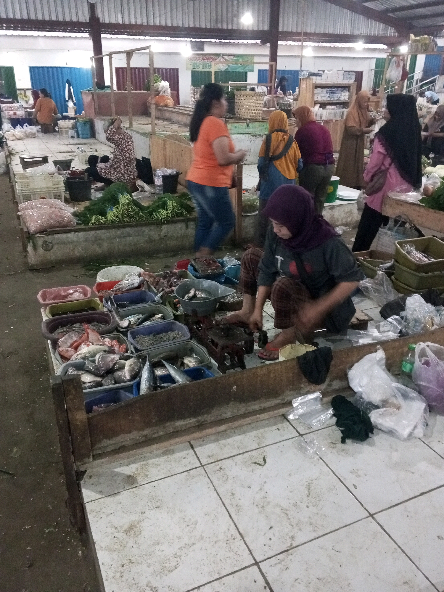 The image captures a scene at an outdoor market in Gentan, Indonesia. In the foreground, a woman wearing a purple hijab and plaid pants is crouched on the ground, appearing to be working with something in front of her. Various plastic bags, some containing goods, and other debris are scattered on the tiled floor around her. Behind her, a wooden stall displays numerous containers filled with fresh fish.
Further into the market, several other individuals are visible. A woman in an orange shirt and blue jeans walks past, while others are engaged in shopping or working at their stalls. The market appears to have a covered roof structure supported by metal poles. Stalls are set up with tables and displays, featuring goods such as vegetables and more fish. The overall impression is of a busy, active marketplace with a variety of vendors and shoppers.