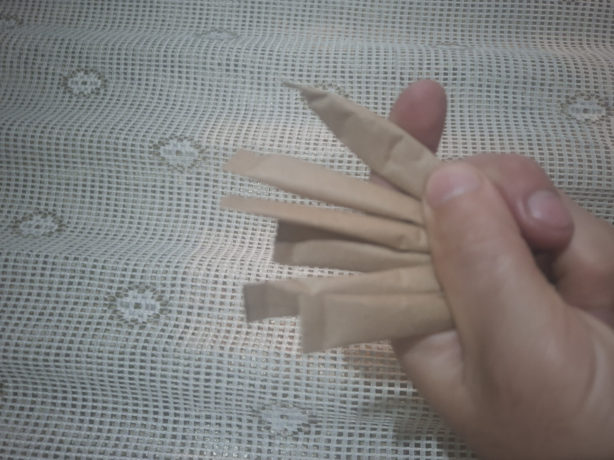 A right hand holds several tan, rectangular, paper-wrapped objects in front of a white, mesh-like surface. The paper objects appear to be small packets or sticks. The hand's fingers are partially visible, gripping the objects. The background consists of a surface with a repeating, grid-like pattern.