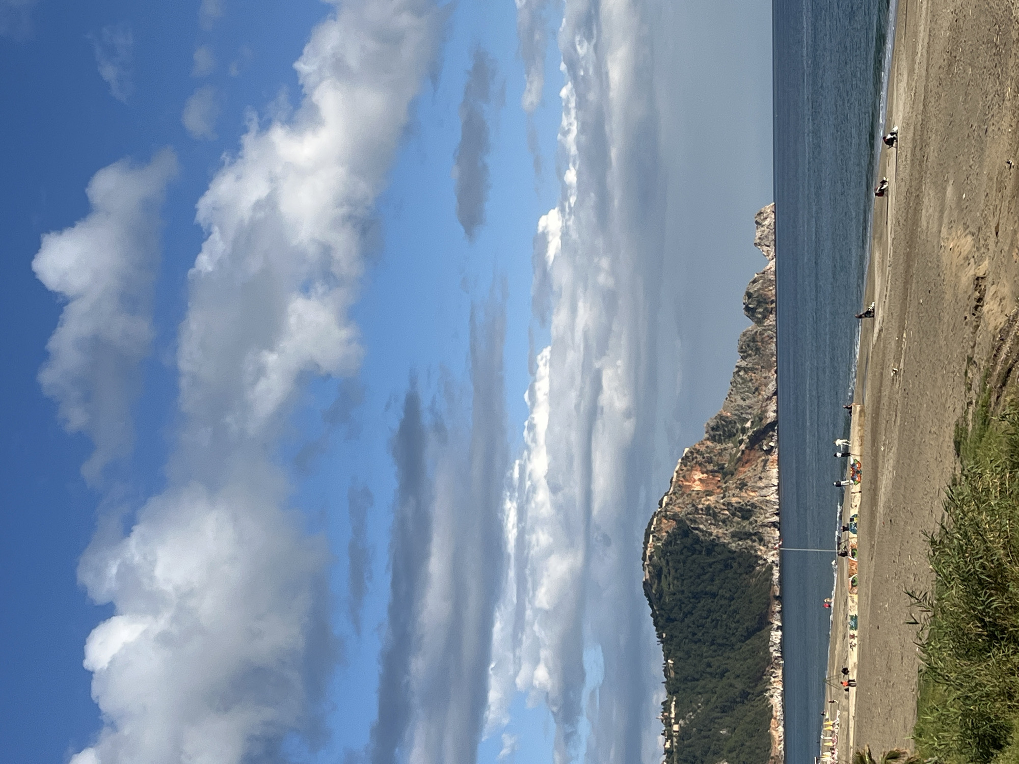The image shows a beach scene in Alanya, Turkey. The sky is blue with white and gray clouds. To the right is a body of water that meets a sandy beach. Along the beach, there are people and what appear to be beach umbrellas. A large rocky hill covered in greenery is next to the beach and water. The photo appears to be taken from an elevated position, looking down towards the beach. A few green bushes are visible at the bottom right corner of the image.