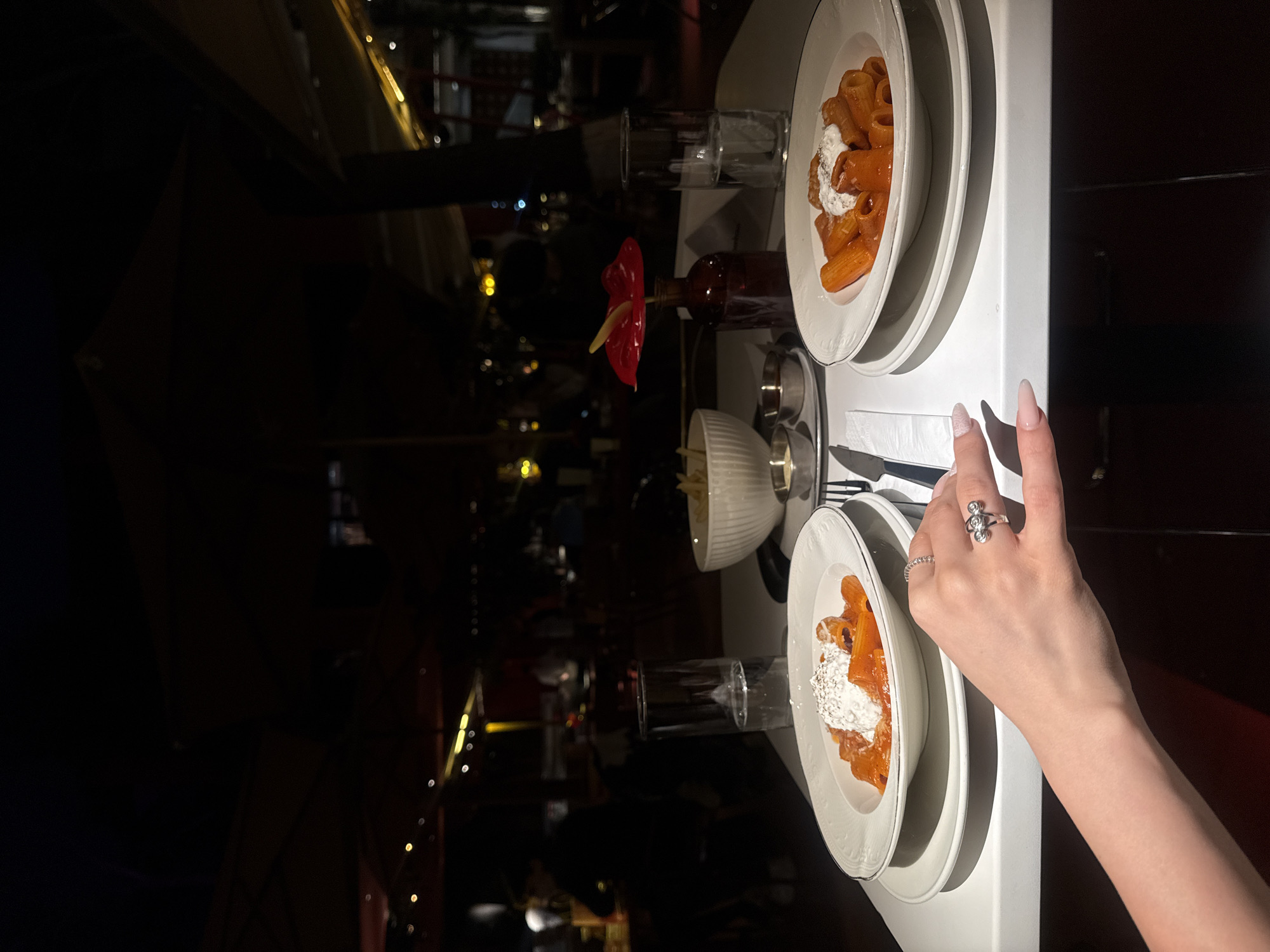 The image shows a dinner setting at a restaurant in Yerevan, Armenia. Two plates of pasta with a red sauce and a dollop of white cheese on top are placed on a white table. A small bowl of french fries, cutlery, a glass of water, and a small vase with a single red flower are also present. A woman's hand with rings is partially visible near one of the plates. The background is dark and blurred, suggesting a dimly lit restaurant interior. The scene appears to be a moment captured during a dining experience.