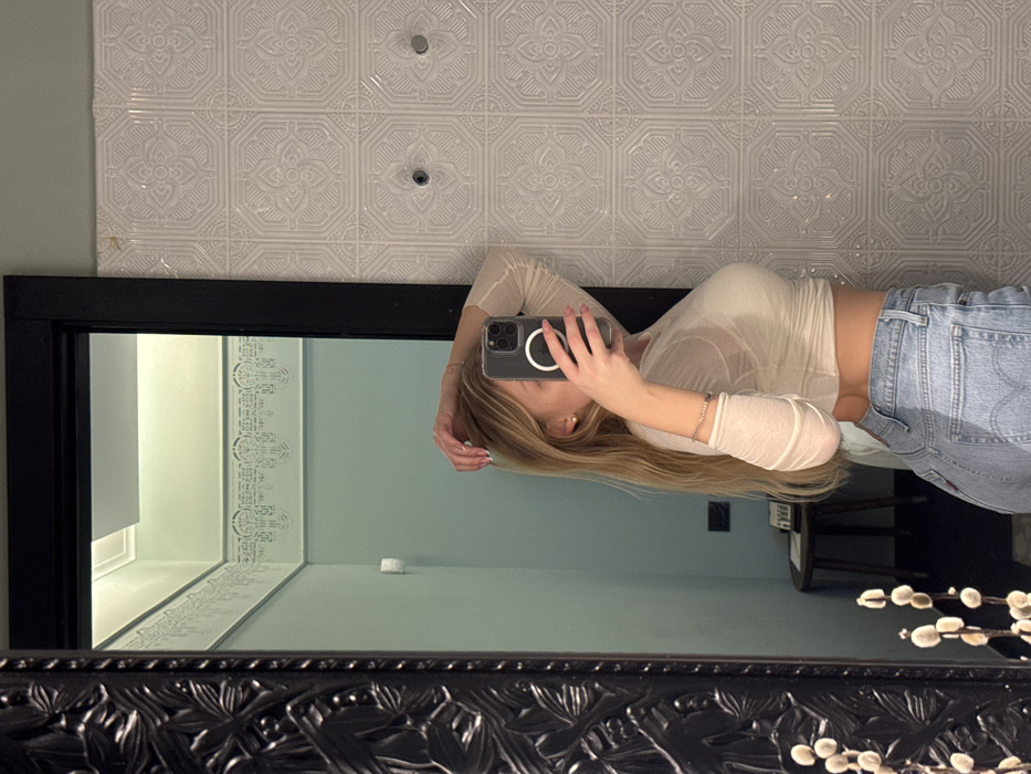 A woman in a sheer, off-white top and light-wash jeans takes a mirror selfie. The mirror has a dark, ornate frame. The reflection shows a blue-toned room with white trim. Above the mirror is patterned white wallpaper with circular accents. A modern iPhone is visible in the woman's hand, partially obscuring her face. The reflection also captures a glimpse of a dark wooden table or stool. The foreground of the mirror features decorative branches with white buds.