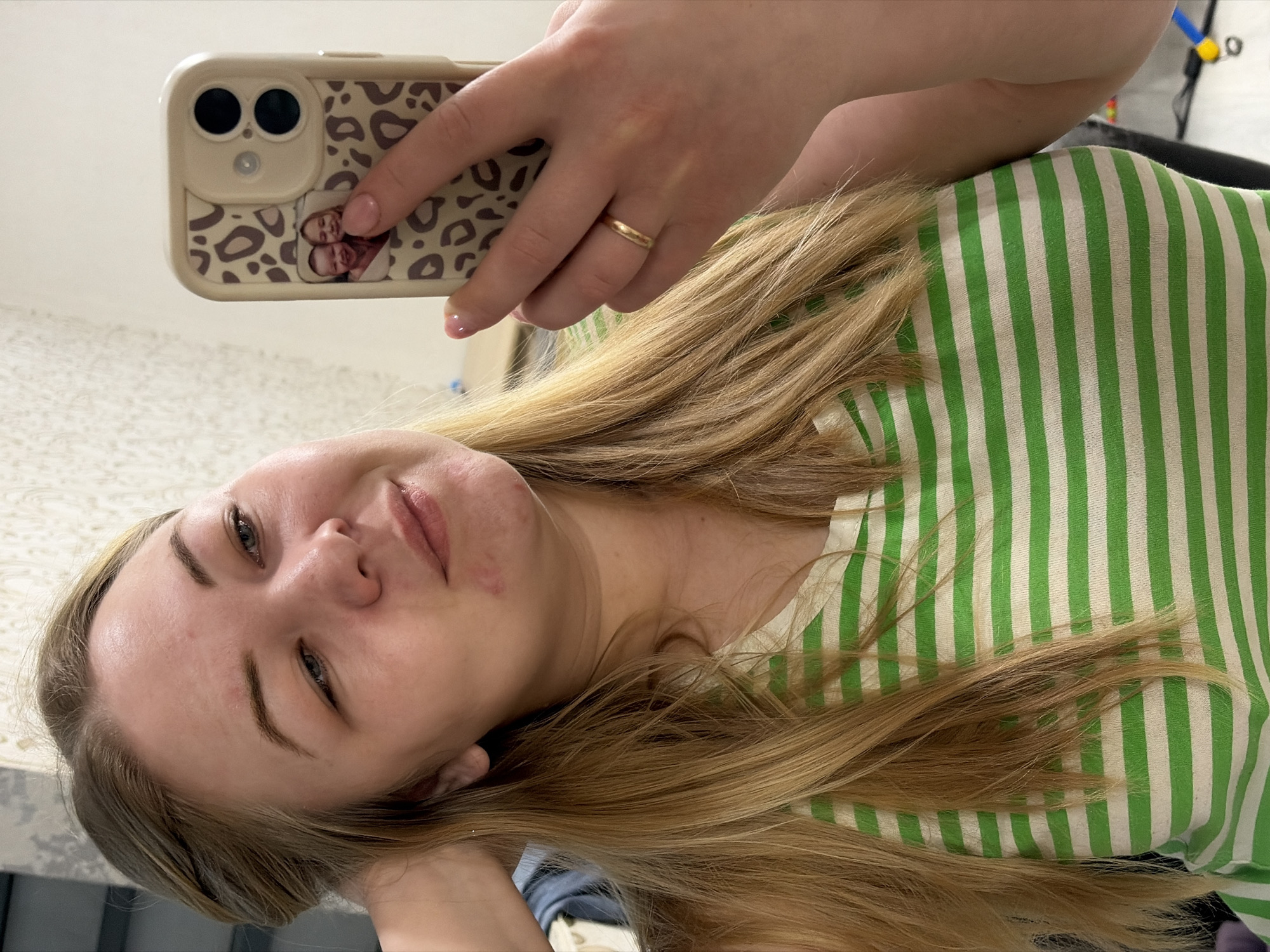 A fair-skinned woman is taking a selfie with a smartphone. The phone has a beige case with a leopard print and a small photo attached. The woman is wearing a gold ring on her left ring finger. She has light brown hair and is wearing a green and white striped shirt. The background appears to be an interior space. The location in Noviy, Russia is not discernible from the image.
