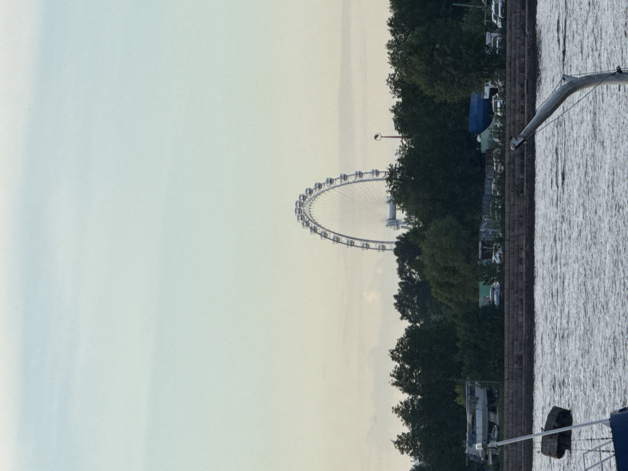 The image, originally rotated 90 degrees clockwise, displays a waterfront scene in Saint Petersburg, Russia, likely during the early morning or late afternoon given the soft, muted light.
In the middle-left of the horizontally-corrected frame, a large Ferris wheel dominates the skyline, its white structure and gondolas visible against a pale, slightly hazy sky. To the right of the Ferris wheel, a dense line of lush green trees runs along a waterway. Visible through and above these trees are various indistinct structures, including what appear to be parked vehicles, possibly small buildings, and a distinctive blue tent-like roof. A street lamp is also noticeable among the trees.
To the right of the trees, a dark, brick-like embankment separates the land from a body of water, possibly a river or canal. The water's surface is textured with ripples, reflecting the ambient light. In the immediate foreground on the right, parts of a boat are visible, including a prominent silver-grey mast or boom extending diagonally across the frame, connected to rigging. Blue railings and a section of a blue boat cover are also seen at the very bottom right.
The overall atmosphere is calm and serene, with no specific human activity clearly depicted. The lighting suggests an overcast sky or diffused sunlight. No discernible text is visible within the image.