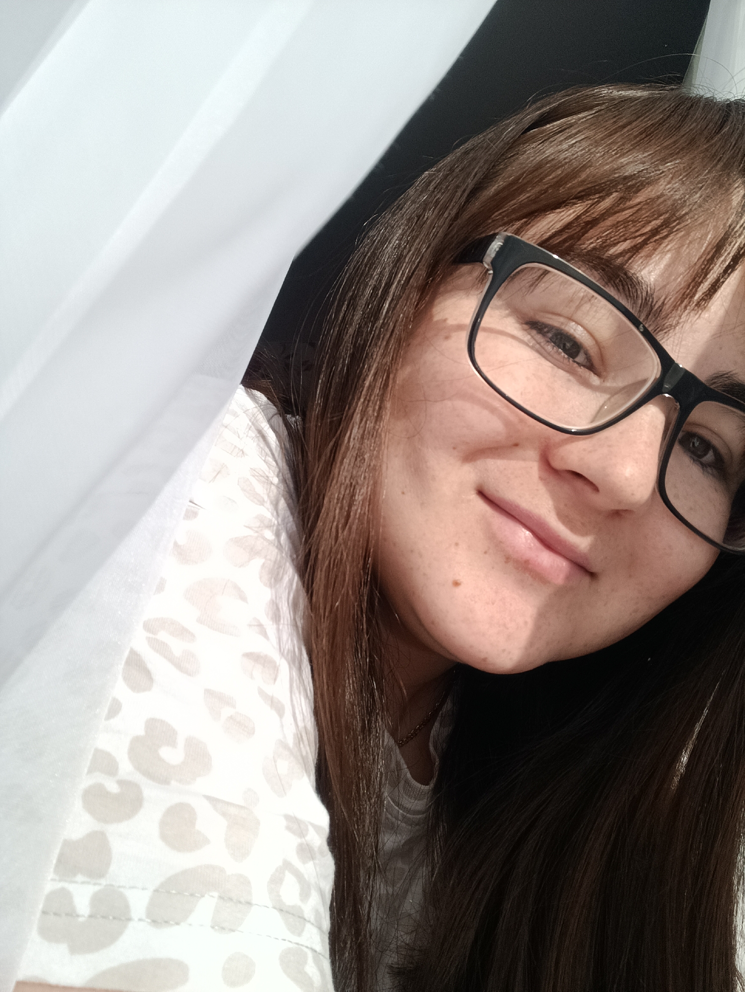 A close-up indoor shot shows a woman with brown hair and bangs, wearing black-framed glasses, and a light-colored shirt with a beige pattern. She is partially obscured by a white curtain on the left side of the frame. Her face is turned towards the camera with a slight smile. The lighting appears natural, suggesting a daytime setting. There is no indication of the city Noviy, Russia.