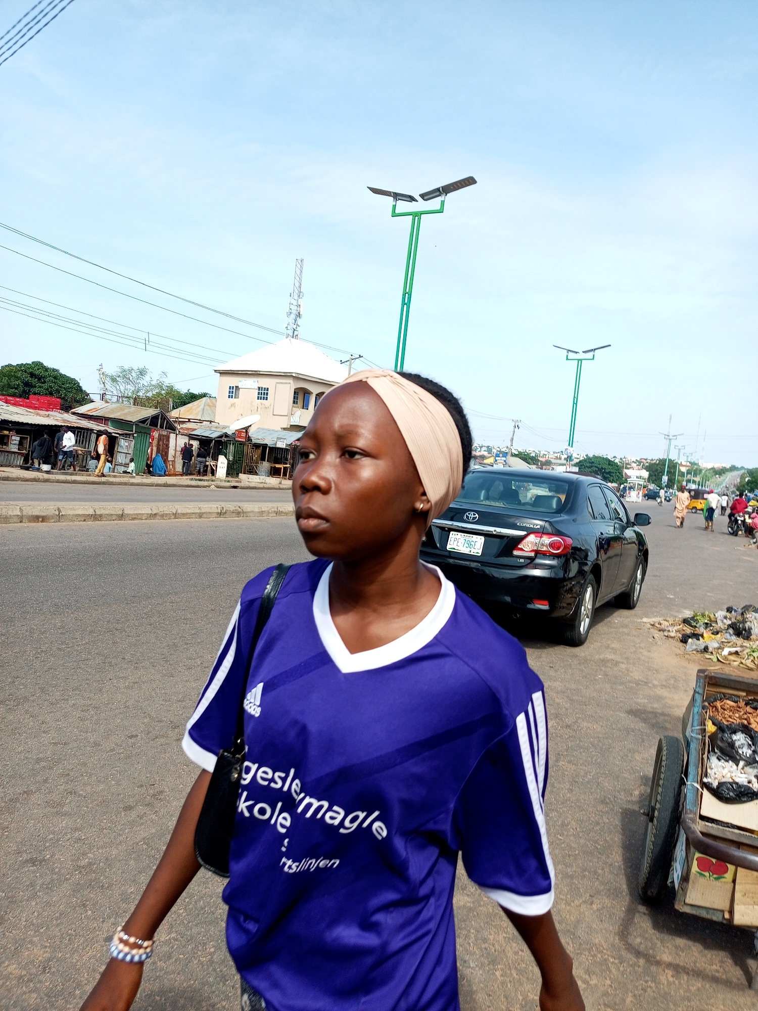 A young woman with a beige headscarf walks on the side of a paved road in Tempure, Nigeria. She is wearing a purple Adidas jersey with white stripes on the sleeves. The woman is looking to her right, away from the camera. Behind her, there is a black Toyota Corolla with a Nigerian license plate. Further back, there are several roadside stalls and buildings, as well as streetlights and power lines. A few other people are visible in the background, walking or standing. A small cart filled with what appears to be trash or discarded items is parked on the right side of the road. The sky is clear and blue.