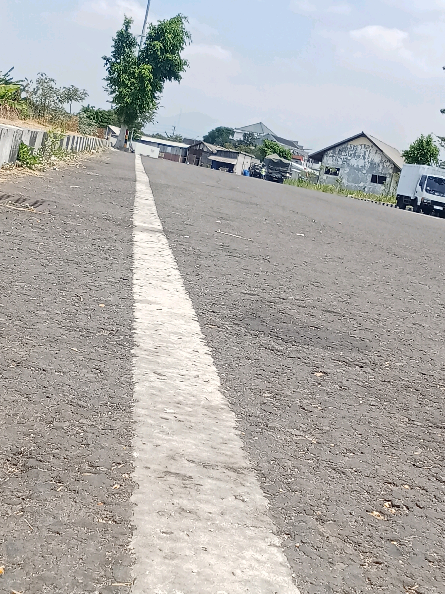 The image depicts an outdoor scene in Sukowinangun, Indonesia, likely a street or access road within an industrial or commercial area. The foreground is dominated by a grey asphalt road with a prominent white dividing line running diagonally from the bottom left to the upper center of the frame. The road surface appears somewhat worn and uneven, with scattered debris like small stones and dry vegetation.
To the left of the road, there is a concrete barrier or curb, beyond which is dry earth and some sparse vegetation. A tall, leafy green tree stands near the curb, casting a slight shadow. Further back, a light pole is visible.
In the background, several buildings are present. These appear to be utilitarian structures, possibly warehouses or workshops, with varying architectural styles and states of repair. Some have pitched roofs, while others are flatter. One building in the mid-ground has a weathered, grey facade with visible signs of wear.
Several vehicles are visible in the background. A white truck, possibly a delivery vehicle, is parked on the right side of the road. To the left, a green tarp-covered truck or cargo vehicle is parked. Other indistinct vehicles and structures suggest ongoing activity in the area.
The sky is a pale blue with some wispy clouds, indicating a daytime scene under fair weather conditions. The overall impression is one of an industrial or logistical hub, with a functional and somewhat unkempt appearance. There are no people clearly visible in the image.