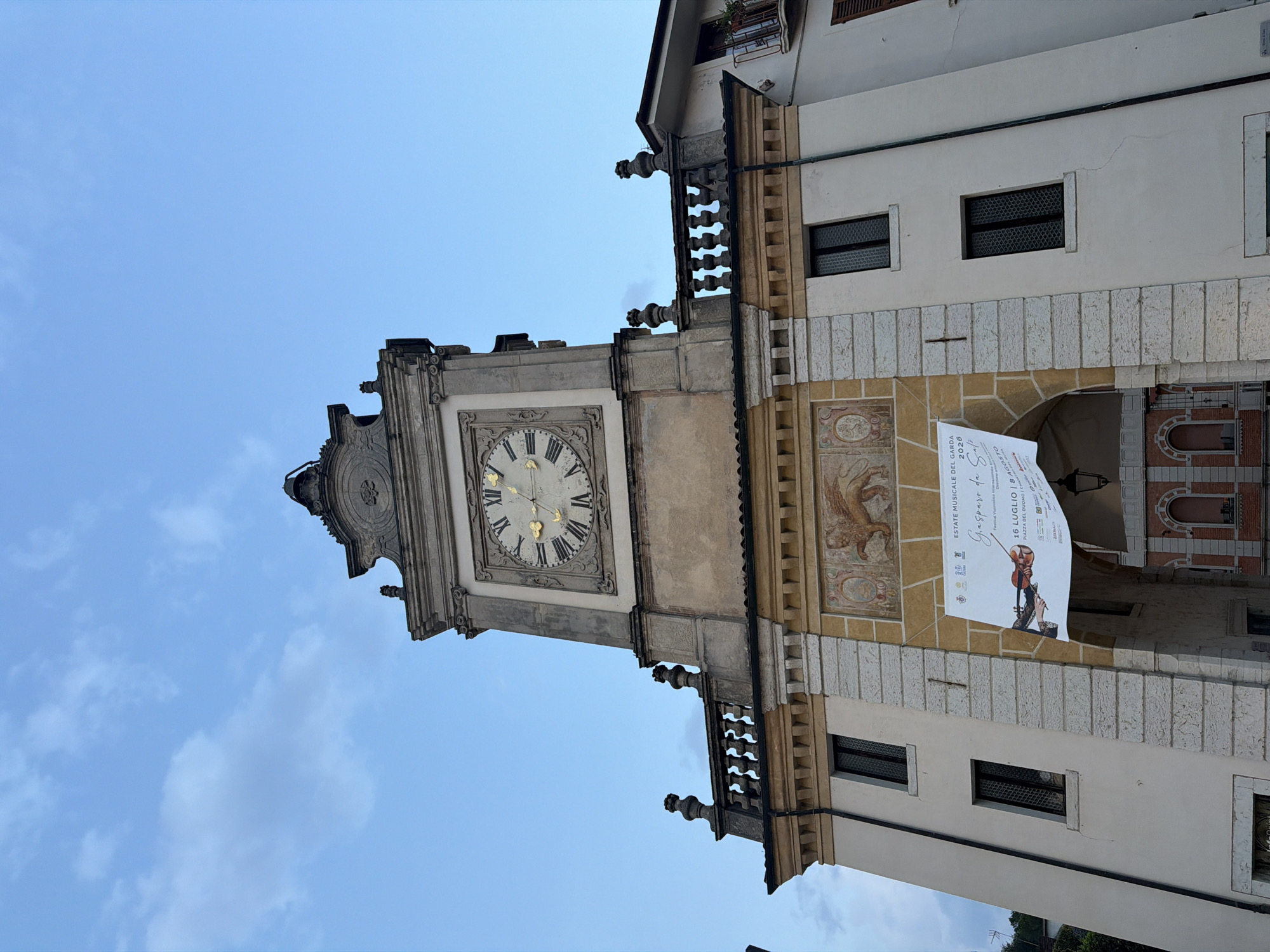 The image shows the Palazzo della Magnifica Terra, located in Salò, Italy, under a cloudy sky. The facade features a large clock with Roman numerals and golden hands, topped by decorative elements. Below the clock, there is an archway decorated with a fresco of a winged lion. A banner advertising the "Estate Musicale del Garda 2020" (Garda Musical Summer 2020) is hung over the archway, displaying an image of a hand playing a violin. The building is made of stone and plaster, with several windows visible on its white walls. To the right of the Palazzo, a portion of a modern building with balconies is visible. The lighting suggests it is either early morning or late afternoon.