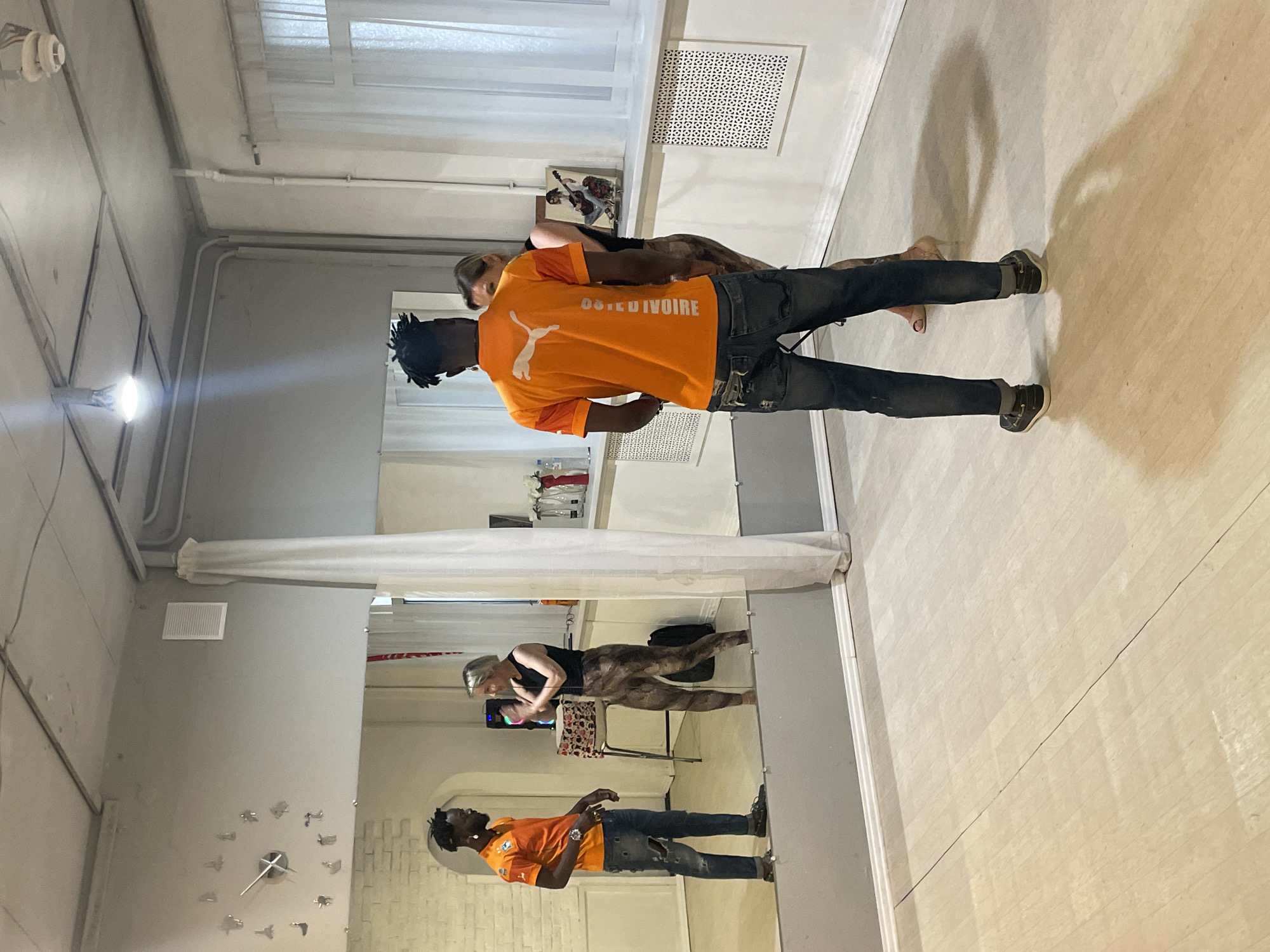 A full shot, rotated, shows a person wearing an orange shirt that says "COTE D'IVOIRE" standing in a room with a mirror. The person has dark skin and dreadlocks. The person is also wearing jeans and dark-colored shoes. In the background, another person wearing dark clothes stands in front of what appears to be a television and some exercise equipment. The room appears to be a dance studio or a gym, given the mirror and the exercise equipment. On the wall, there is a large clock. The picture was taken indoors in Sochi, Russia.