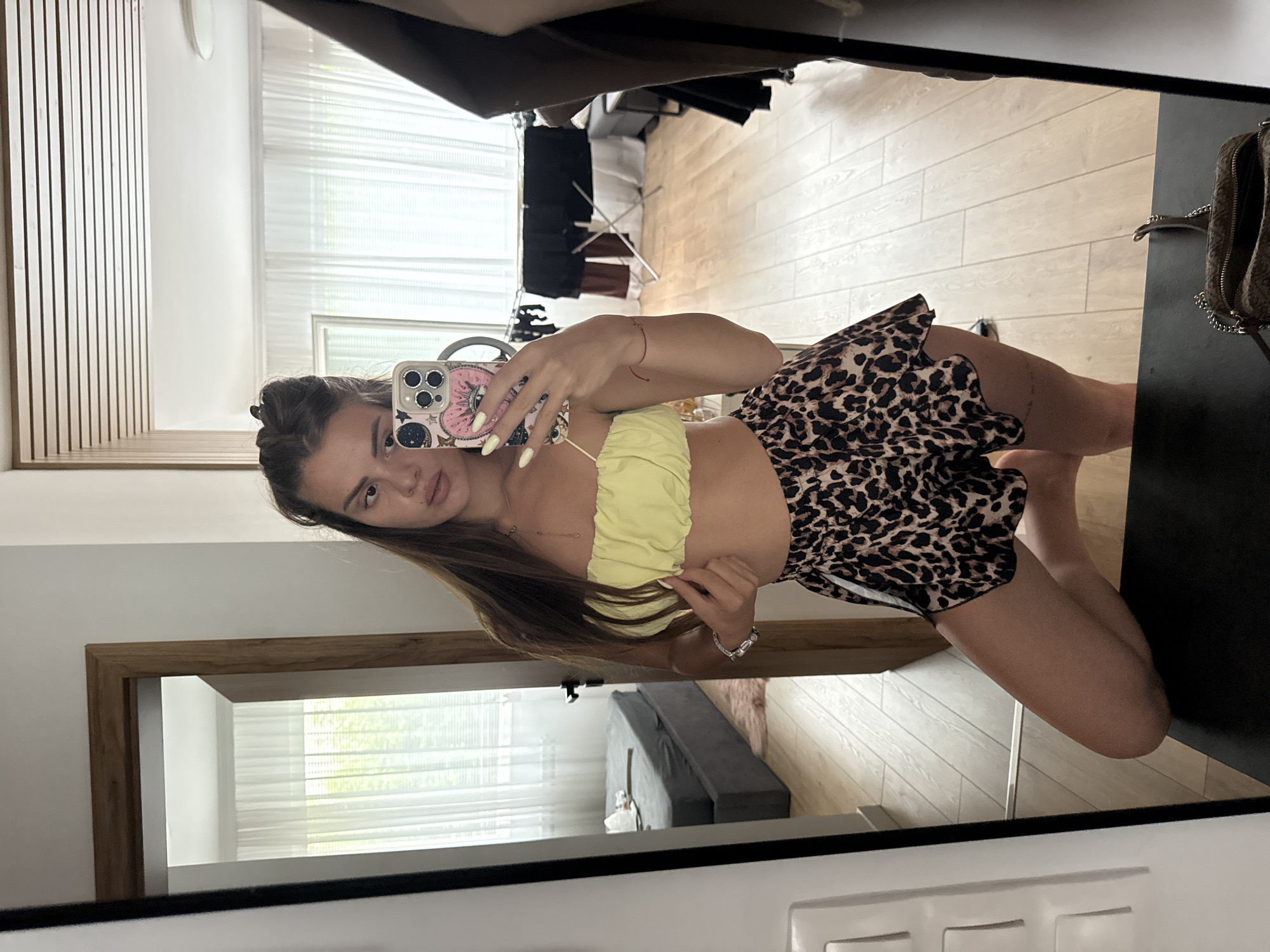 A woman is taking a mirror selfie in what appears to be a bedroom. She is wearing a yellow crop top and leopard print shorts. Her hair is pulled up in a bun. The room has light wood flooring and sheer white curtains. In the background, a clothes drying rack is visible, along with a bed and a doorway. The scene is set in Plovdiv, Bulgaria.