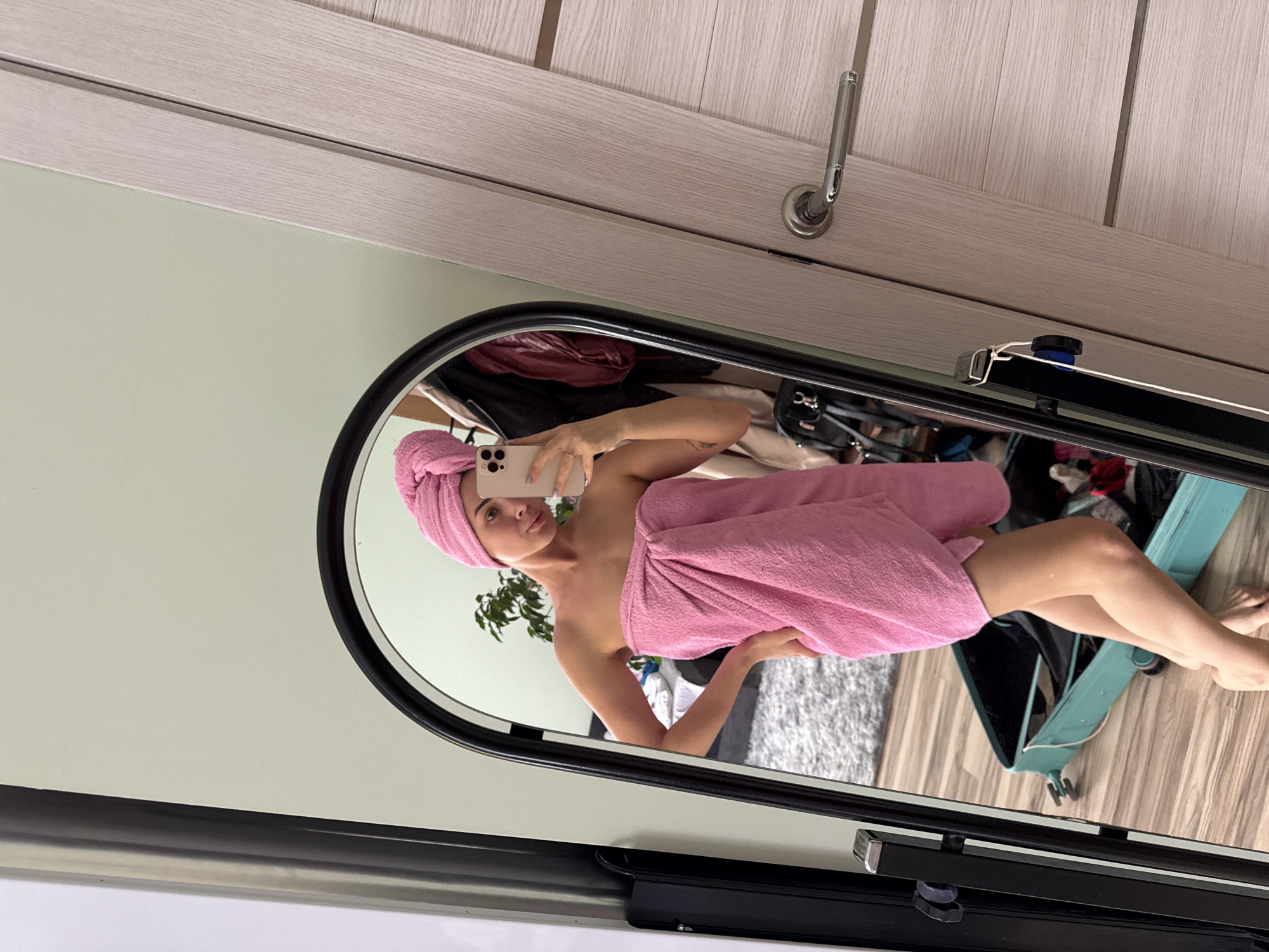 A woman wrapped in a pink towel and a pink towel turban is taking a selfie in a mirror. She is holding a smartphone in her left hand. The reflection in the mirror shows a cluttered room with a plant, clothing, and a blue suitcase. The location is not identifiable as Sarapul, Russia based on visual data.