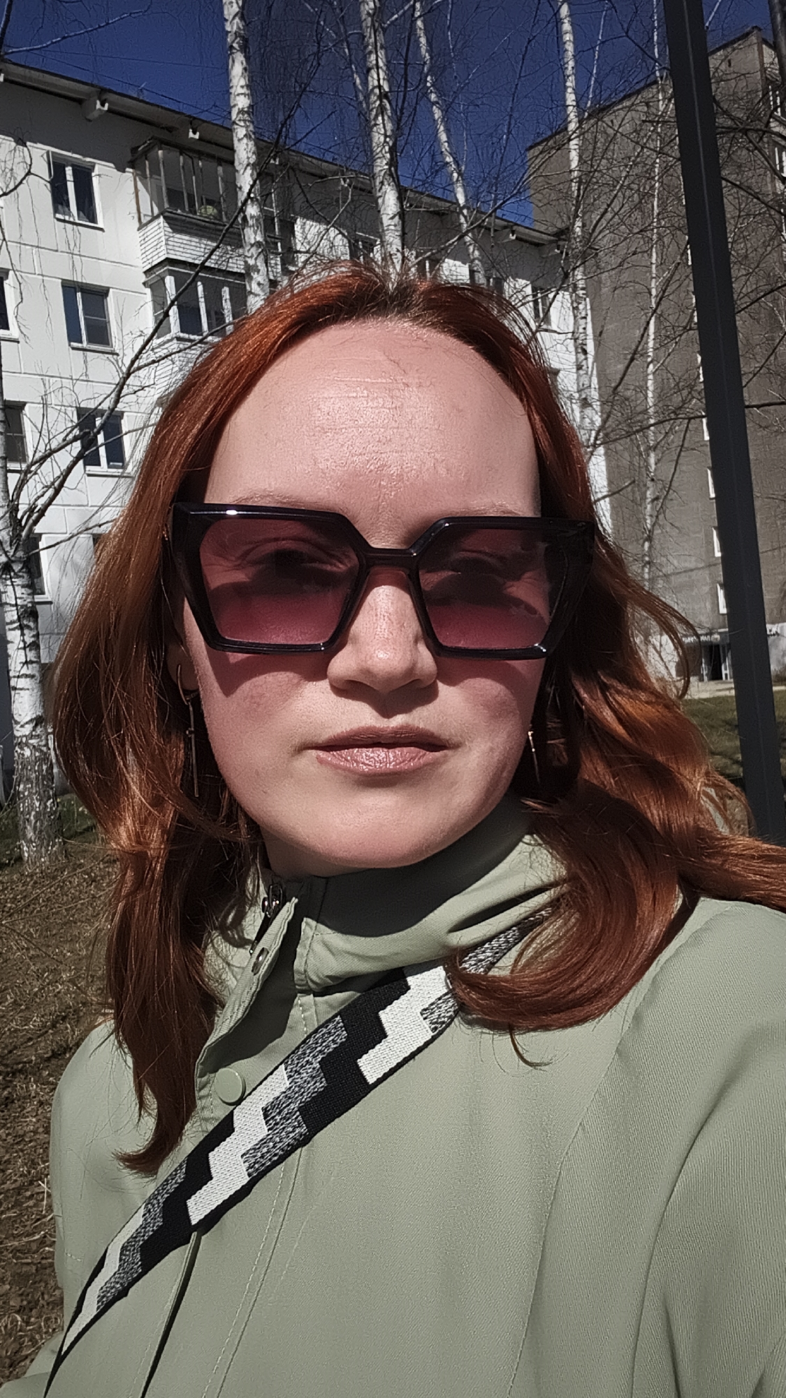 A red-haired woman wearing dark-framed sunglasses and a light green jacket with a black and white patterned strap poses for a close-up selfie outdoors on a sunny day. Behind her are several apartment buildings and bare trees against a clear blue sky. The location is the city of Chaikovsky, Russia.