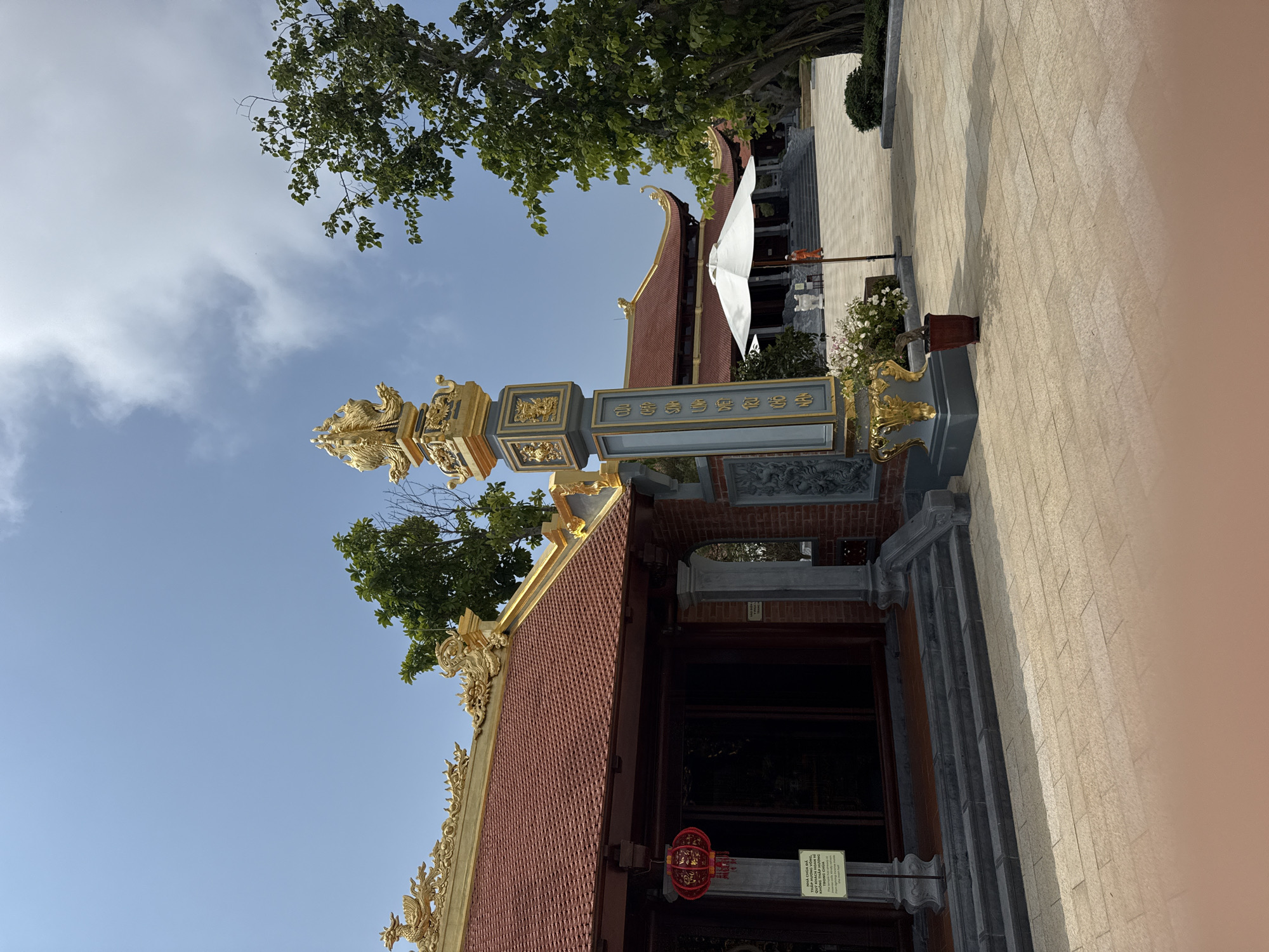 The image depicts an ornate gate or pillar in front of a temple or traditional Vietnamese building. The pillar is decorated with gold accents and a gray-blue central panel with Chinese characters. A red-tiled roof with golden embellishments is visible behind the pillar, and part of the building's entrance is visible with a red lantern and signage hanging on the front. A white umbrella and additional building details can be seen in the background. Above, there is a clear blue sky with some trees. The scene appears to be outdoors on a sunny day. The city is Diên Khánh, Vietnam.