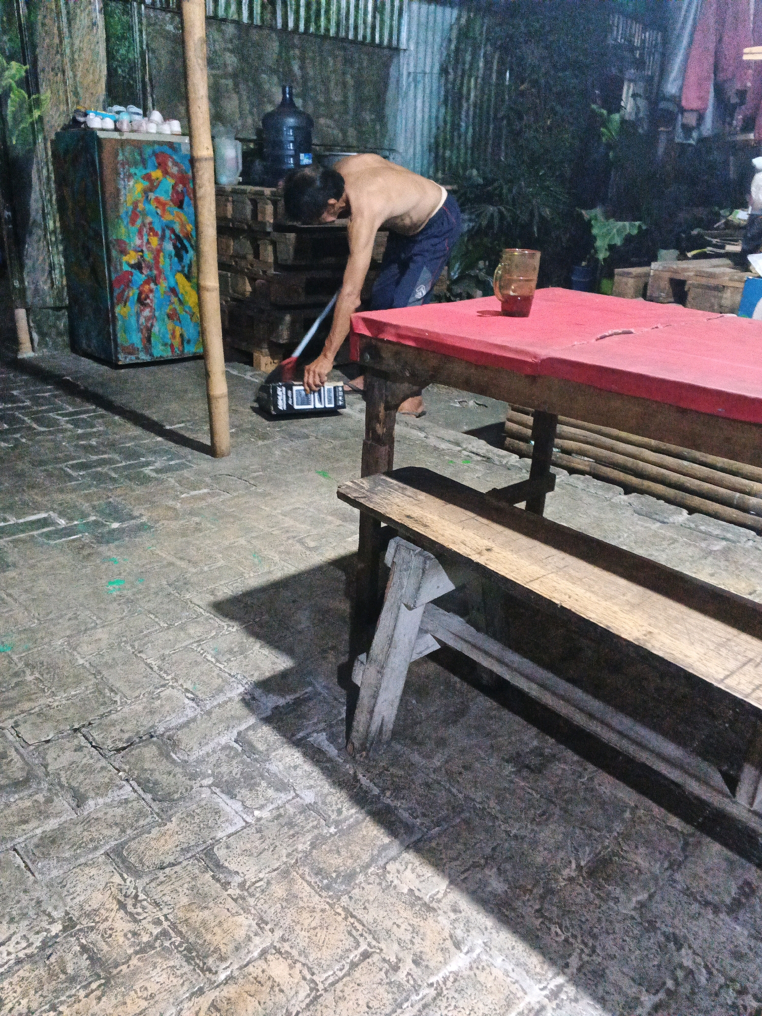 The image shows an outdoor scene in Ungaran, Indonesia. A shirtless man with dark hair is bent over, sweeping the paved ground with a broom. He is wearing dark blue shorts. To his left is a colorful, rectangular cabinet decorated with depictions of koi fish and other aquatic creatures. Behind him, a large blue water jug sits on stacked wooden pallets. In the foreground, a wooden table with a red top and a wooden bench are visible. A glass with a dark liquid sits on the table. The ground is paved with rectangular stones. The background features a corrugated metal wall and lush green foliage. The overall lighting suggests it is evening or nighttime.